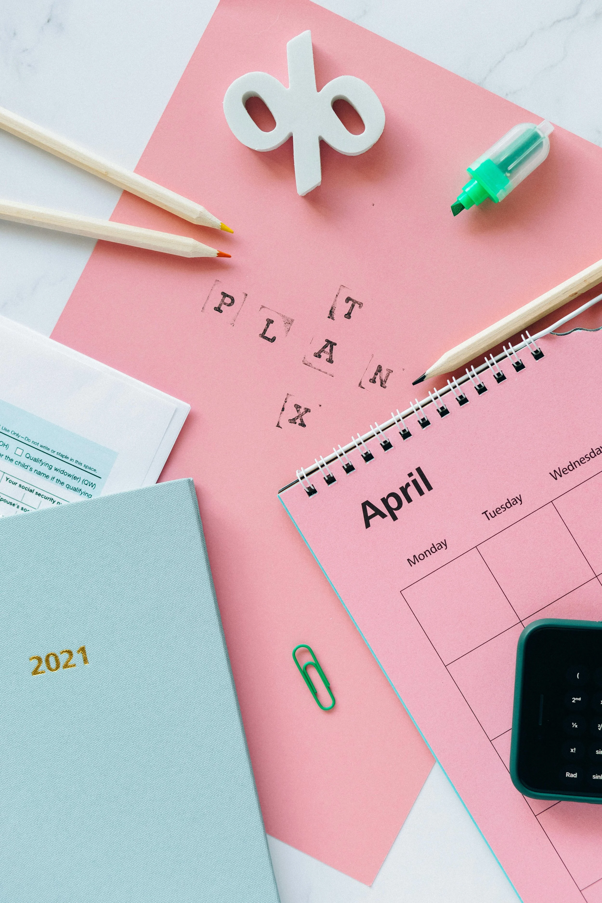 Flat lay of an April calendar on pink paper with two wooden pencils, a green paperclip, a small notebook, a clock, a white decorative symbol, a highlighter, and some documents.