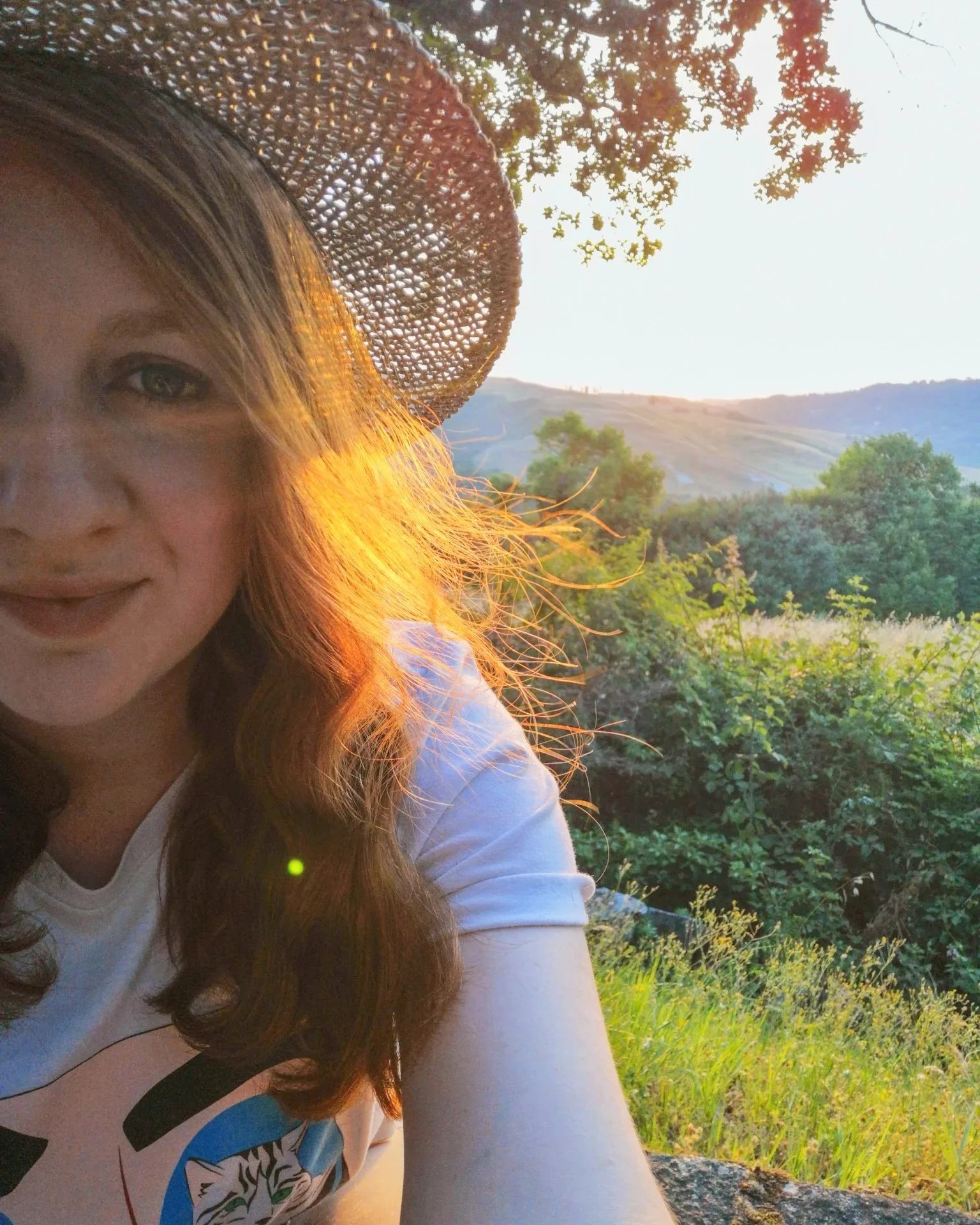 An image of Liv Cassidy, wearing a straw hat outdoors during sunset, surrounded by green trees and hills.