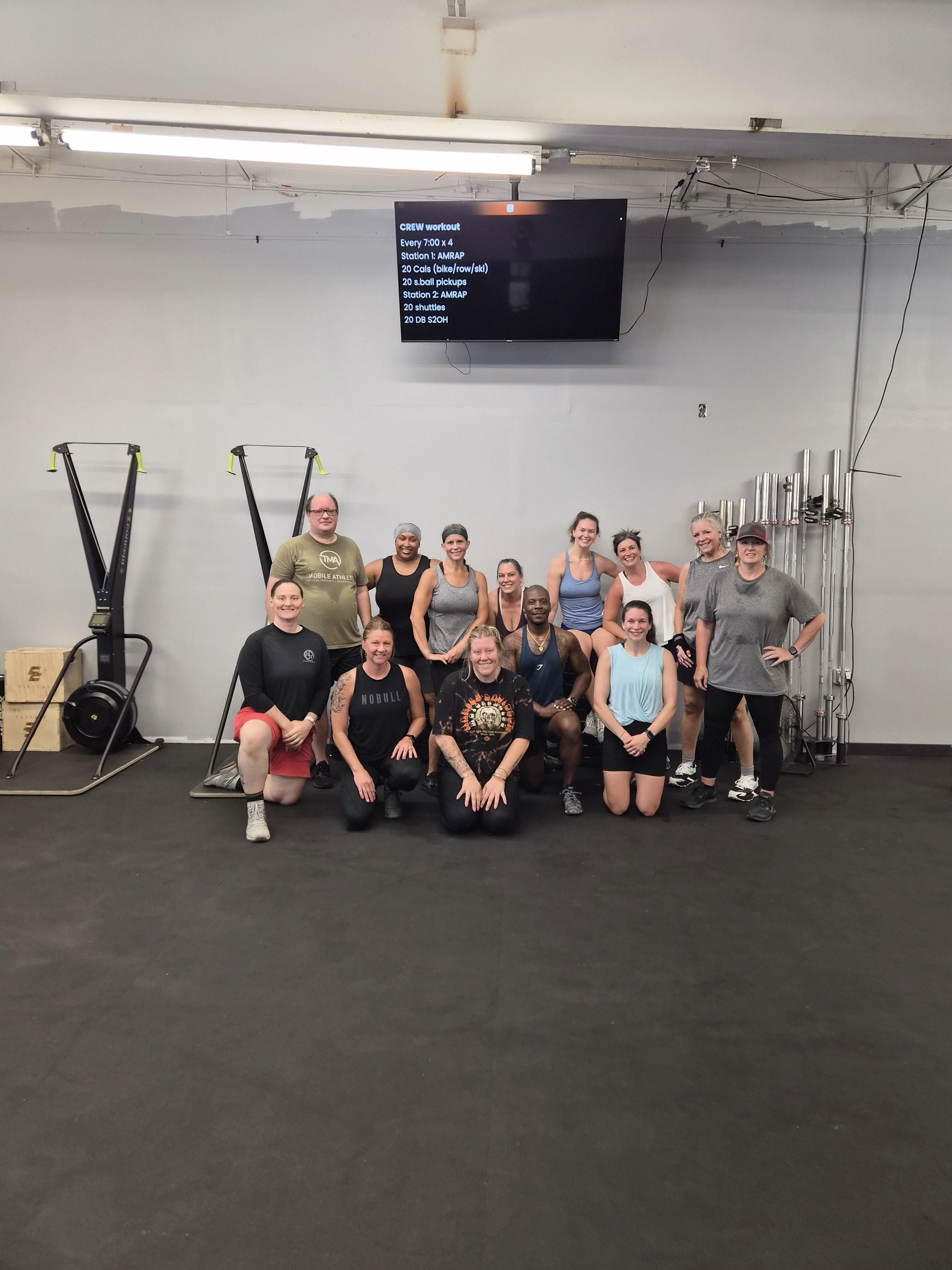 Group of people in workout attire posing for a photo in a gym with workout equipment visible behind them, and a digital screen displaying a workout schedule.