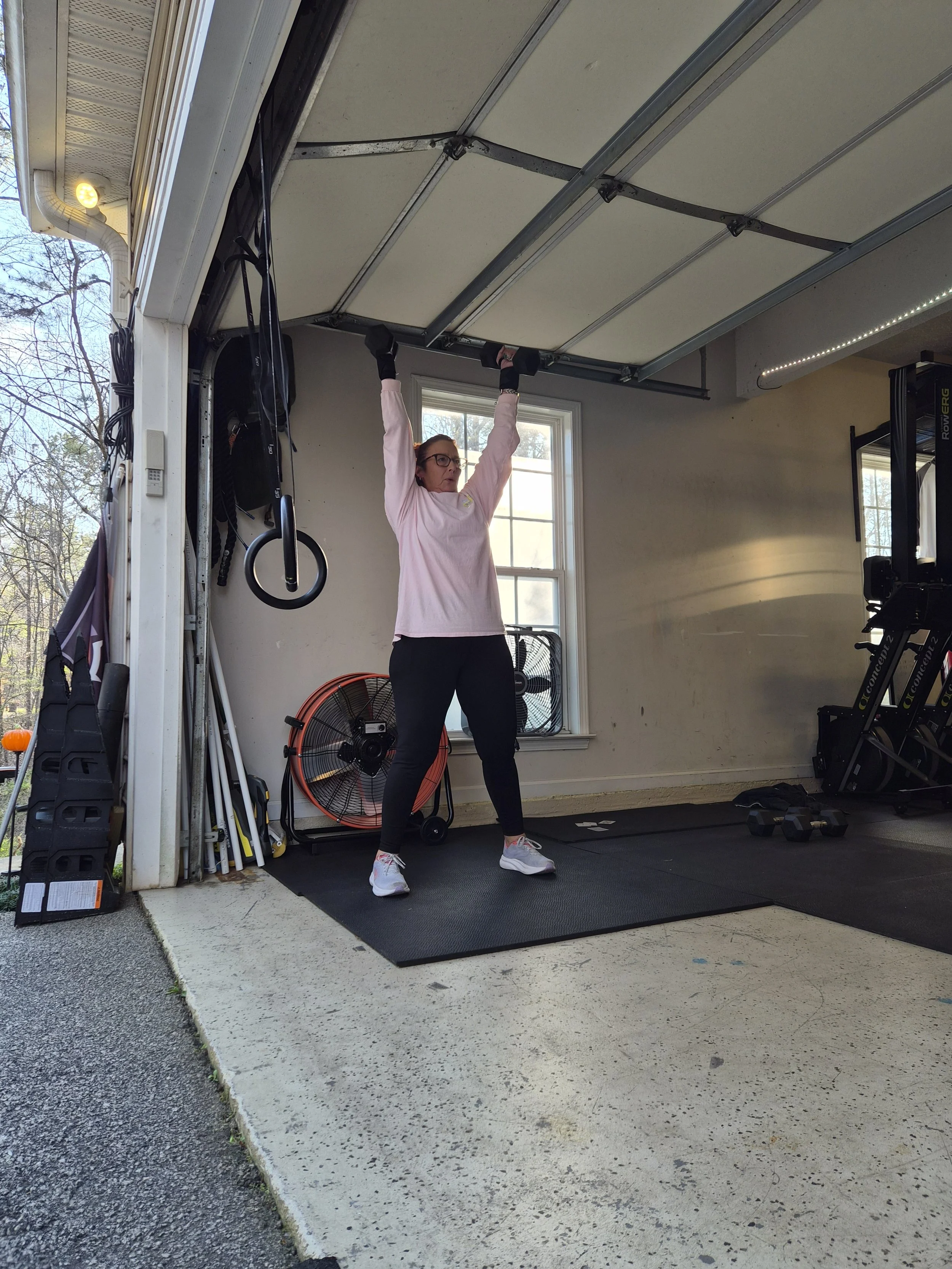 A woman lifting dumbbells in a garage gym, wearing glasses, a pink long-sleeve shirt, black leggings, and white sneakers.