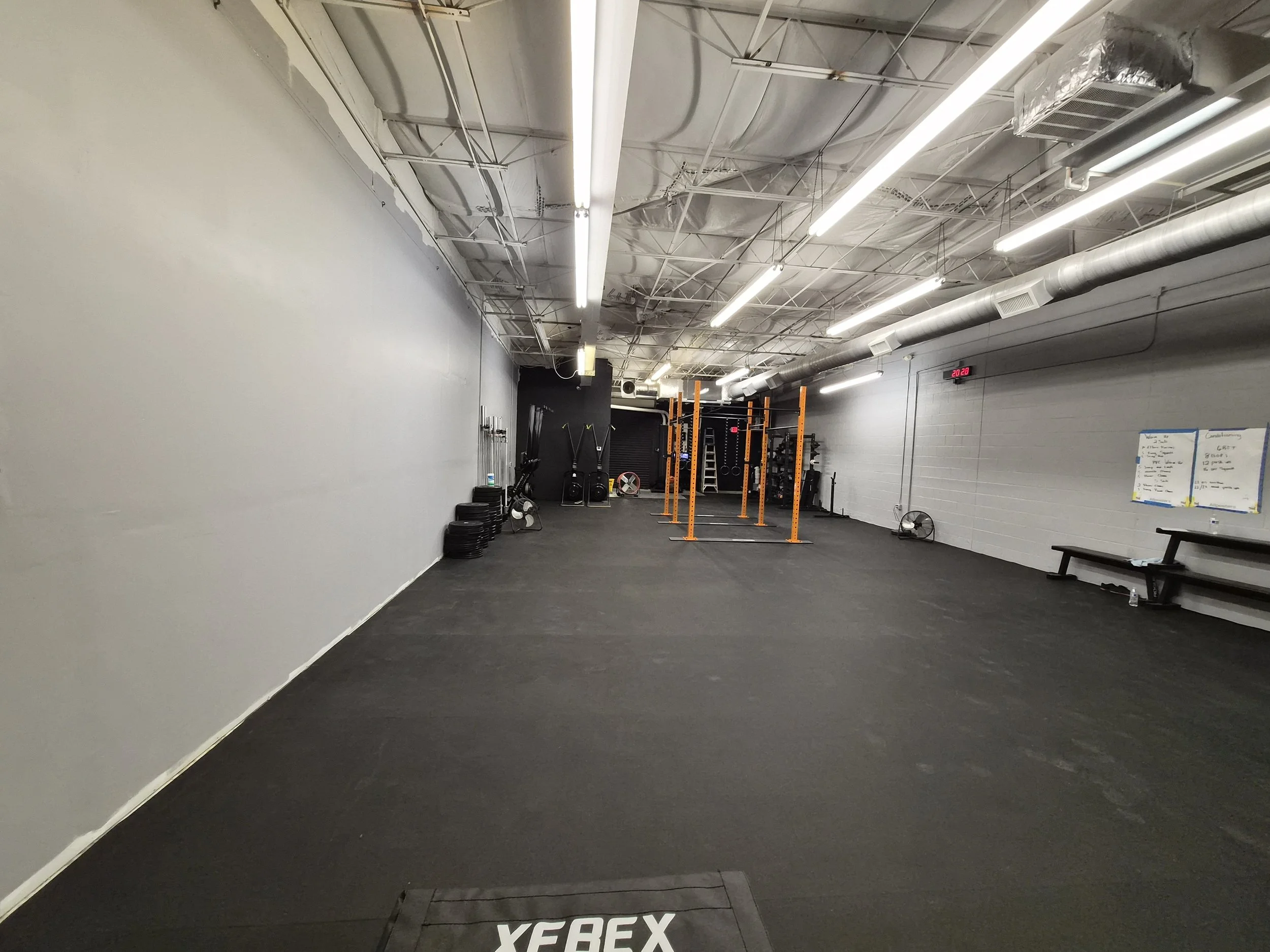 Empty gym with black rubber flooring, gym equipment including pull-up bars and weight plates, white walls, exposed ceiling with lighting, ventilation ducts, and a digital clock on the wall.