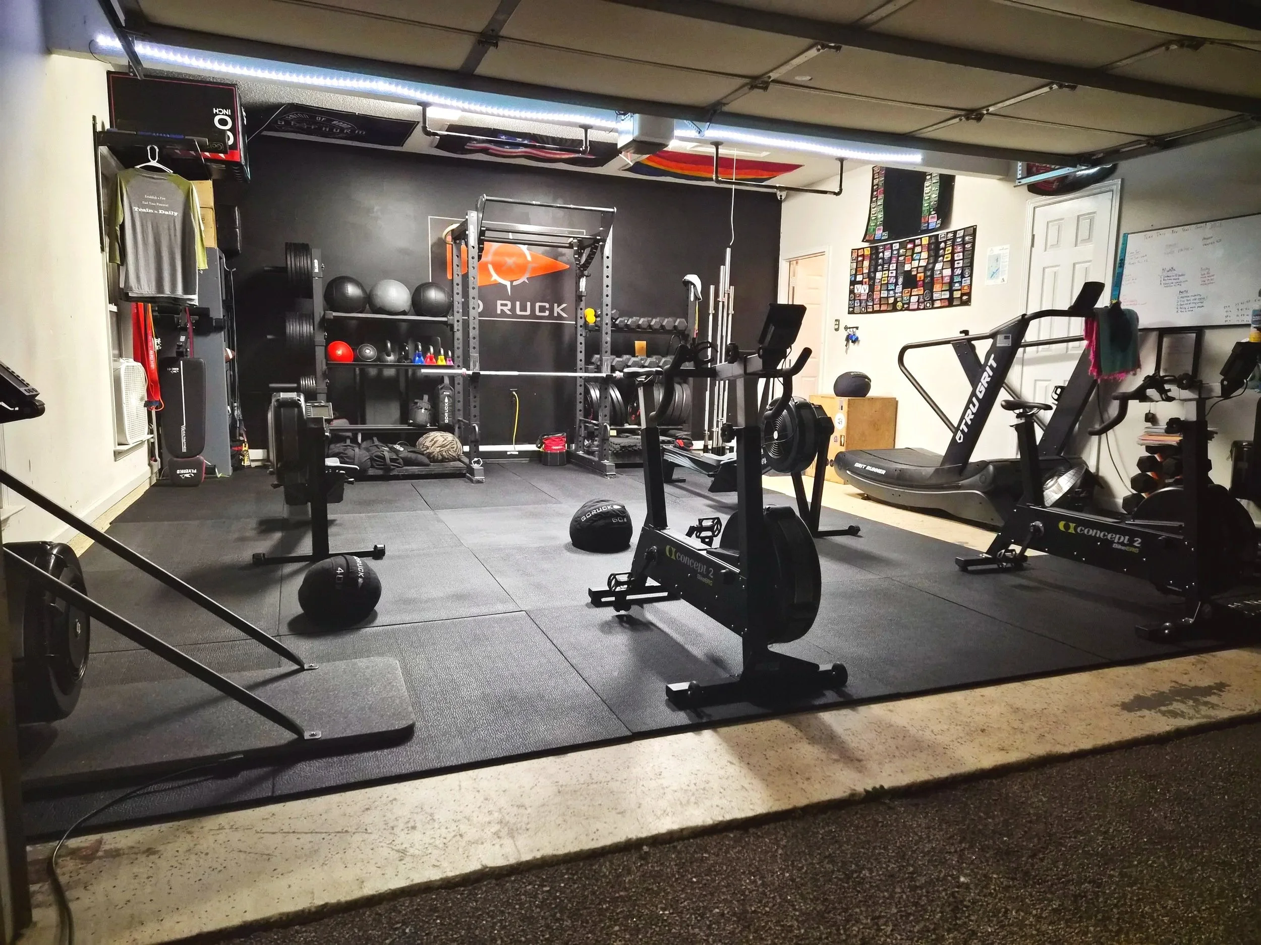 Home gym with workout equipment including a sled, stationary bikes, weight rack, medicine balls, and various exercise accessories in a garage.