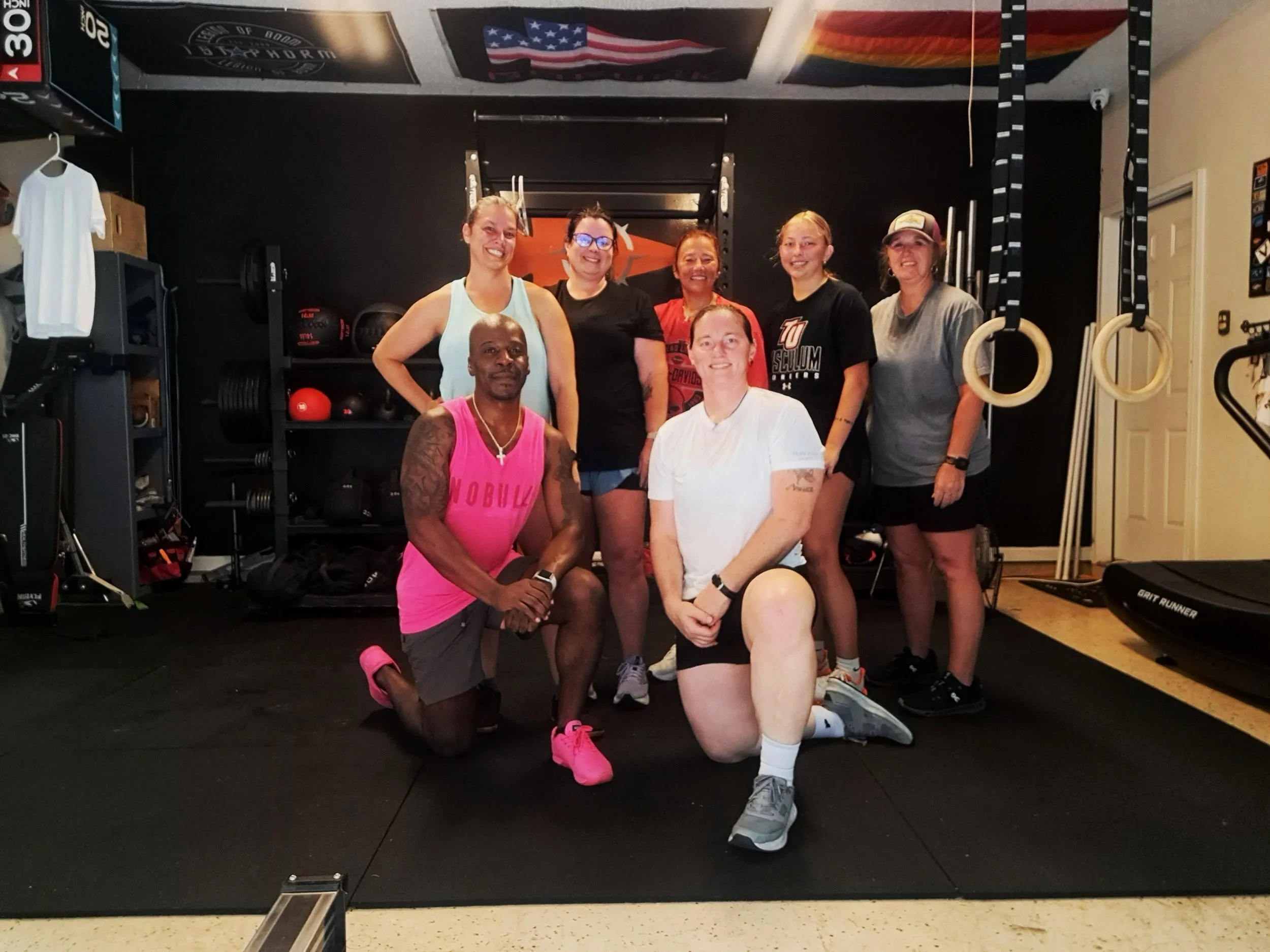 Group of eight women and one man in a gym, posing for a photo. Some are standing, and two are kneeling in front. The gym equipment includes gymnastic rings, medicine balls, weight racks, and a treadmill.