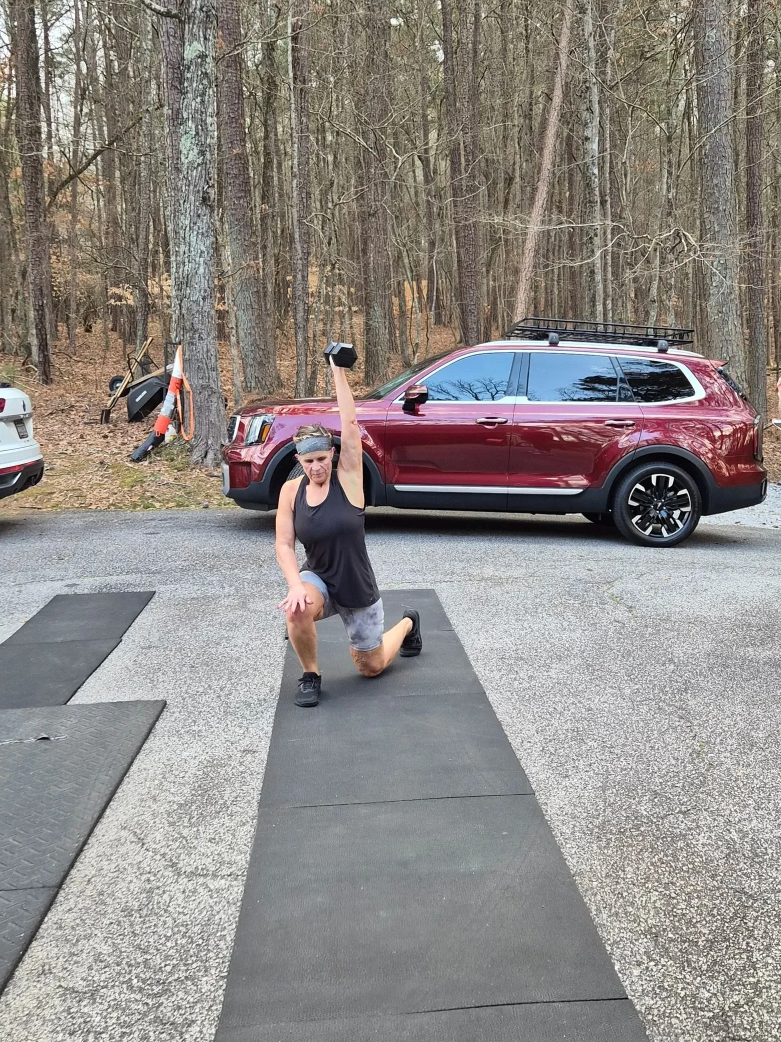 A woman exercising outdoors on a black exercise mat, holding a dumbbell overhead with one knee on the ground, in a wooded area with a red SUV parked nearby.