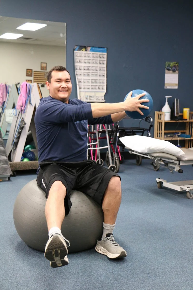 A man smiling while sitting on a stability ball and holding a blue exercise ball in a physical therapy or gym setting.