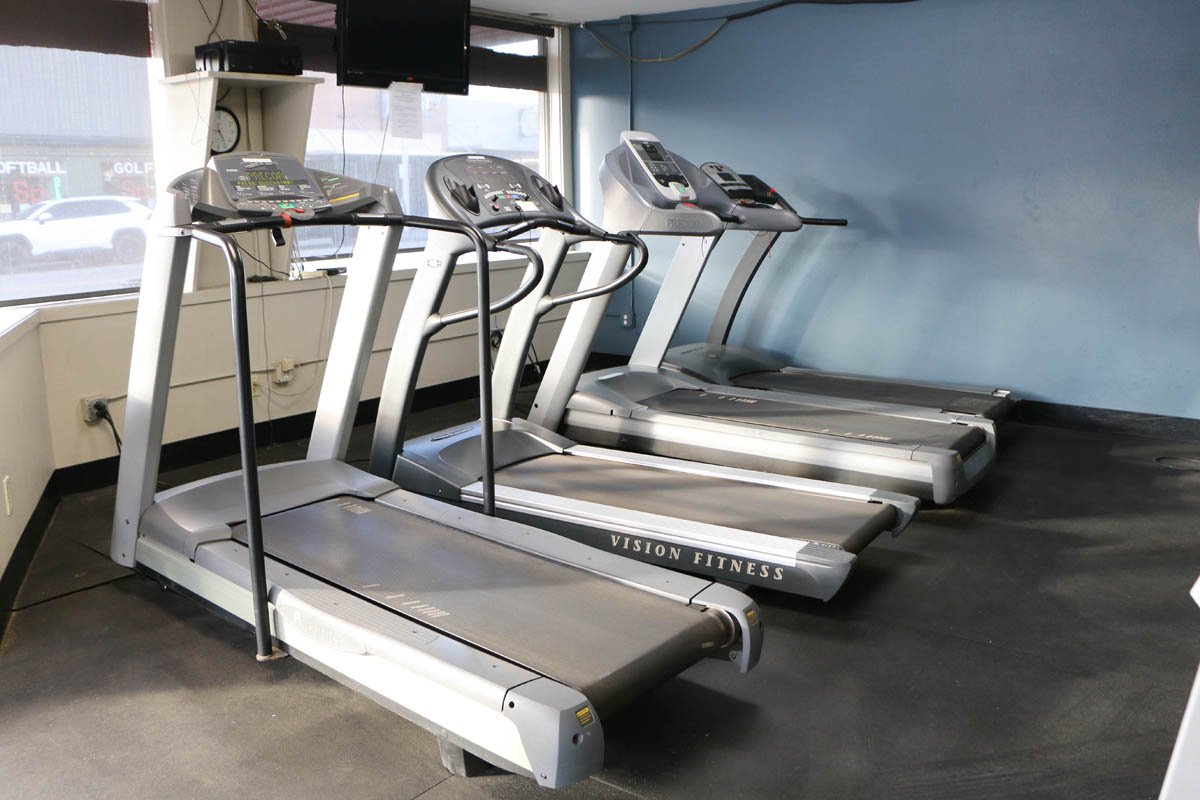 Three treadmill machines lined up side by side in a fitness center, with a window and a TV mounted on the wall behind them.