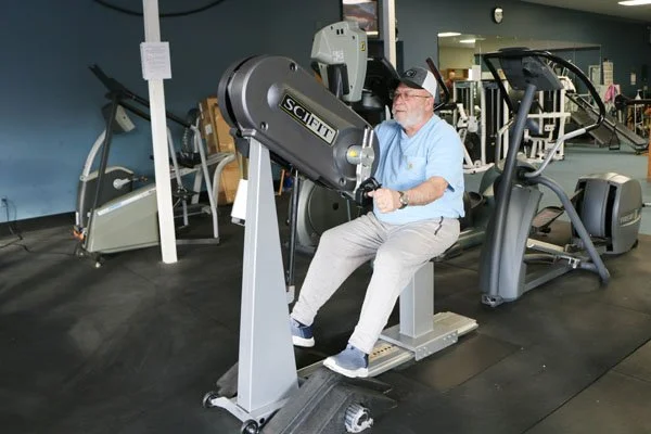 An elderly man using a leg press machine at a fitness gym, with treadmills and elliptical trainers in the background.