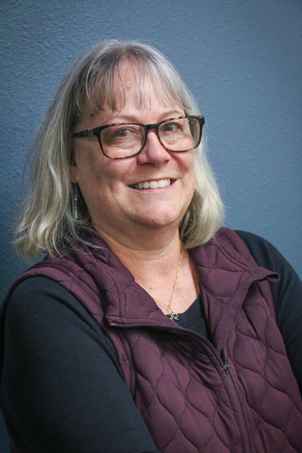 Smiling woman with gray hair, glasses, wearing a black shirt and a purple quilted vest, against a blue wall.