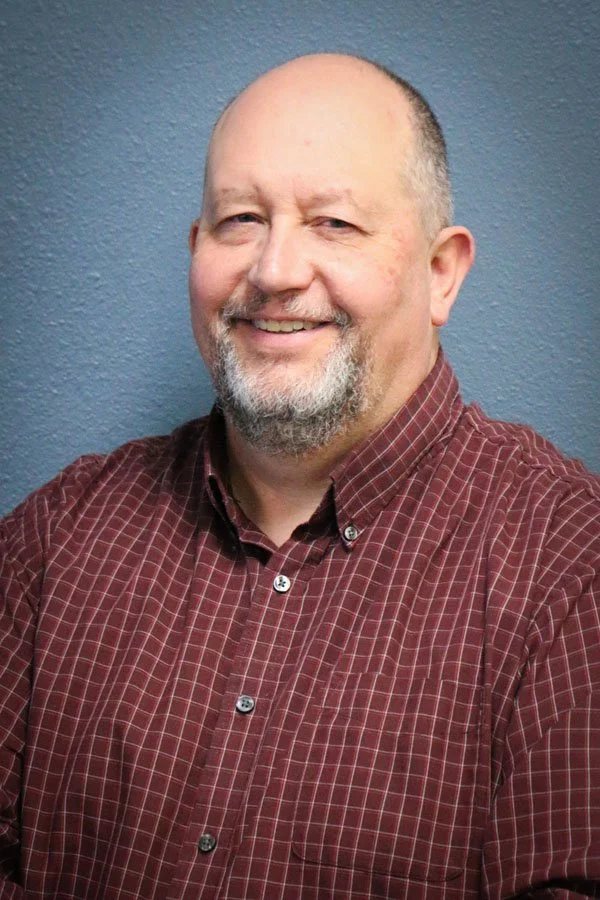 A smiling middle-aged man with a beard and bald head, wearing a red checkered shirt, standing in front of a blue wall.