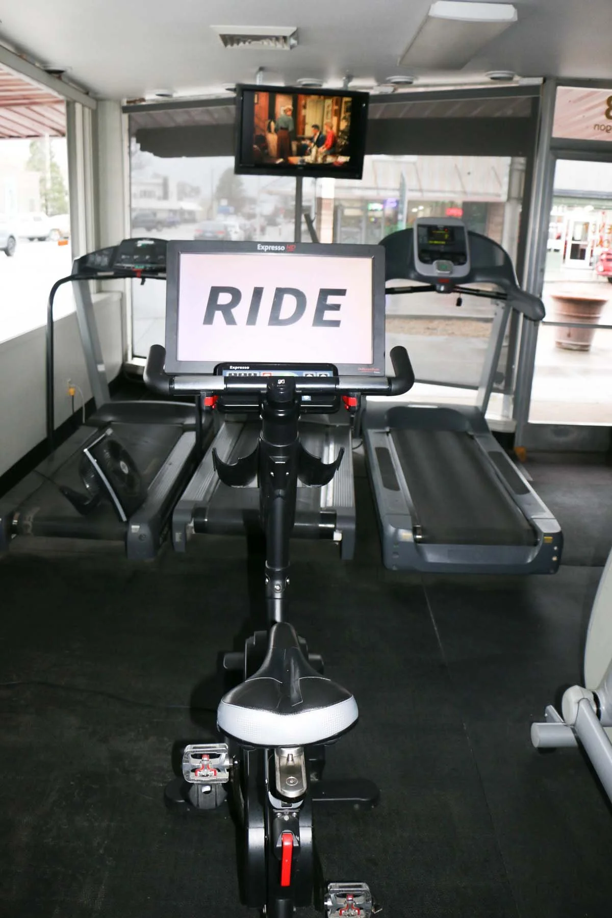 Treadmill on a gym floor with a sign on it displaying the word "RIDE." There is a small monitor screen above showing a scene from a TV show or movie. The background shows windows and part of a parking lot outside.