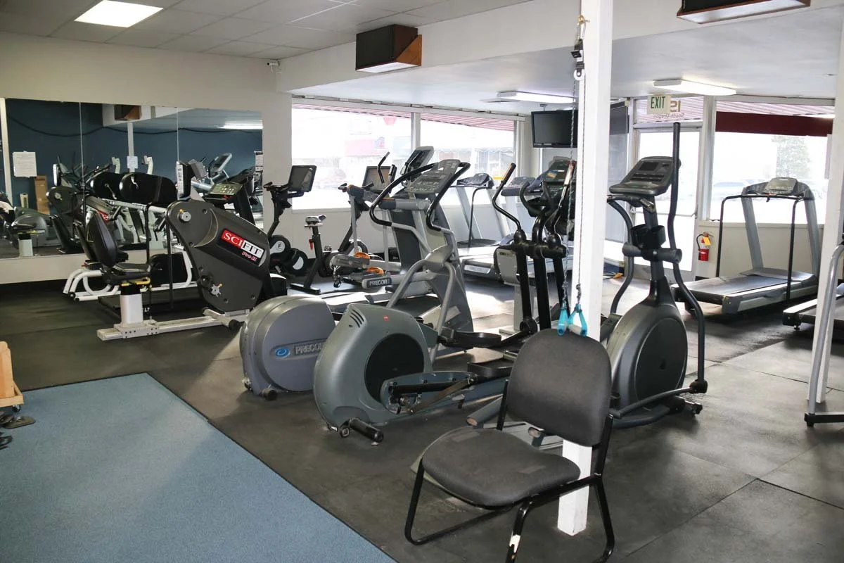 Indoor gym with various exercise equipment including treadmills, stationary bikes, ellipticals, and a chair, with mirrors on the wall and a large window letting in natural light.
