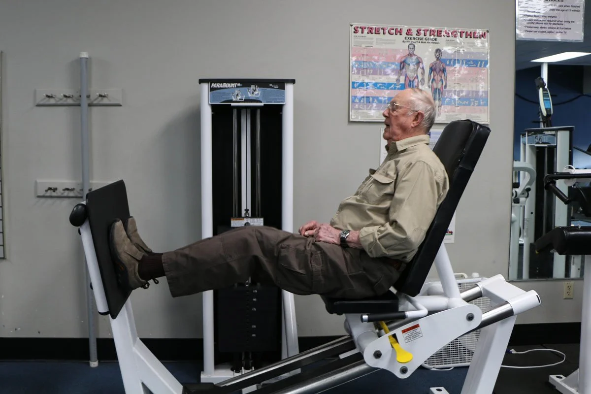 Older man exercising on a leg press machine in a gym, wearing a beige shirt and brown pants.