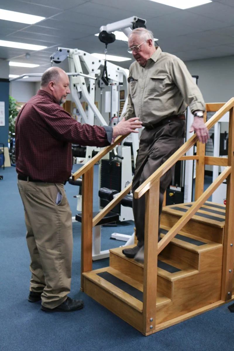 An elderly man using a stair lift device to descend a wooden staircase in a gym or rehabilitation center, assisted by a staff member.