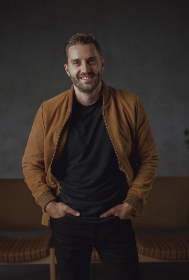A man with short brown hair and a beard standing indoors with hands in his pockets, wearing a black t-shirt and a brown bomber jacket, smiling at the camera.