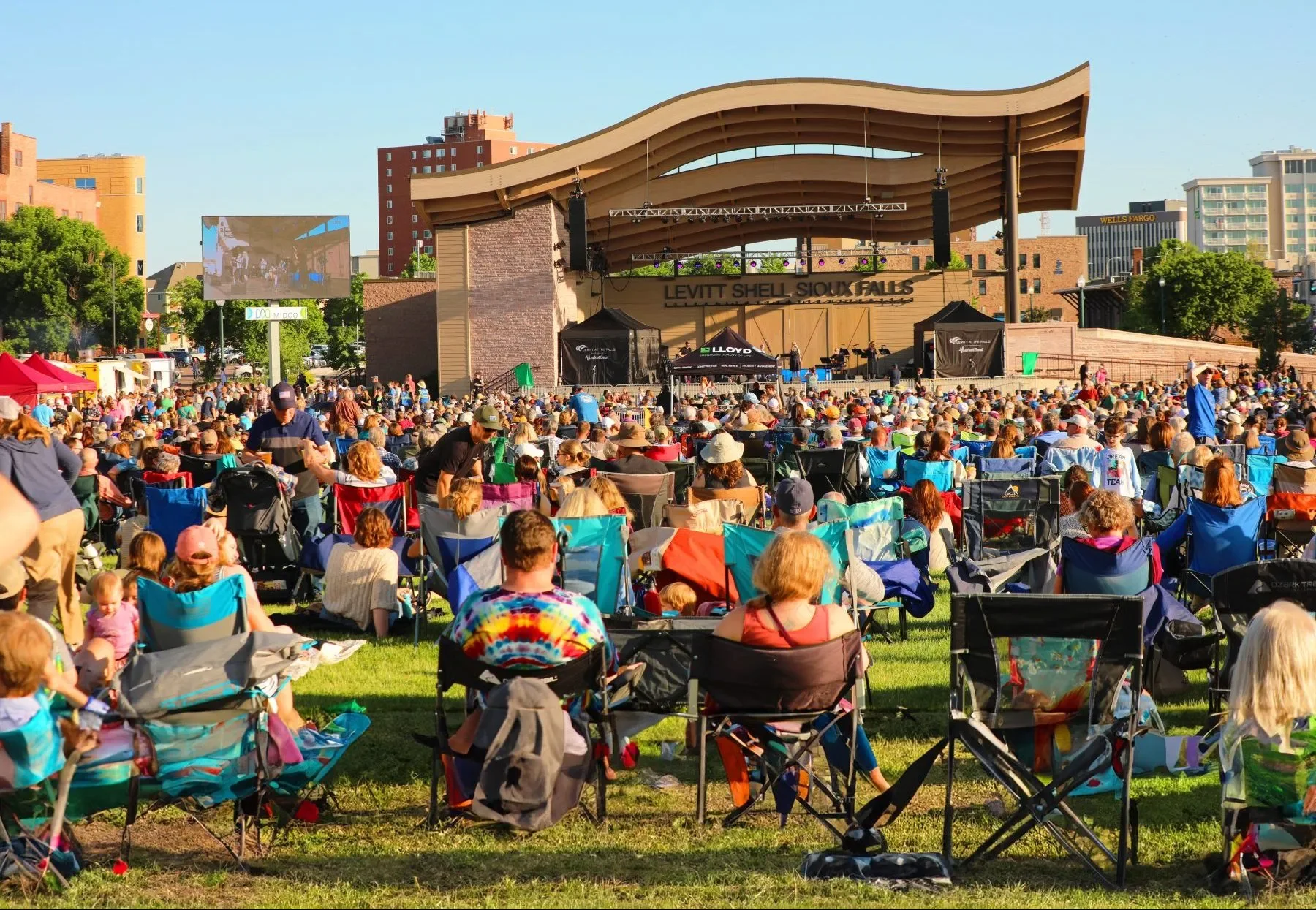 People seated on lawn chairs attending an outdoor concert in Sioux Falls, South Dakota, with a stage and large screen, surrounded by city buildings and trees.
