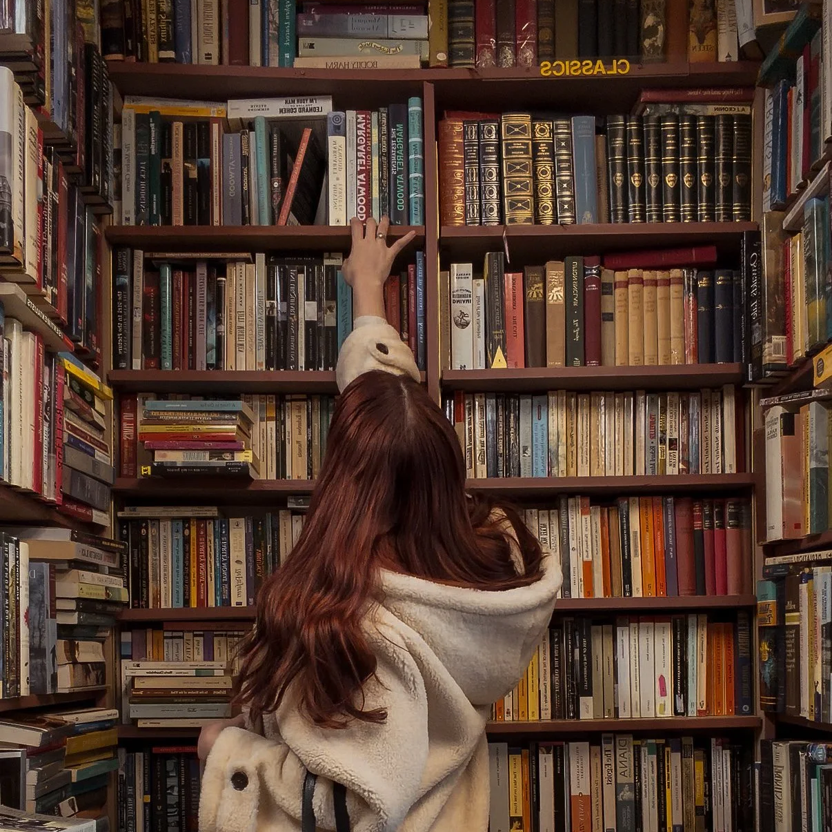 A woman with long red hair reaching up to pick a book from high bookshelf in a bookstore or library.