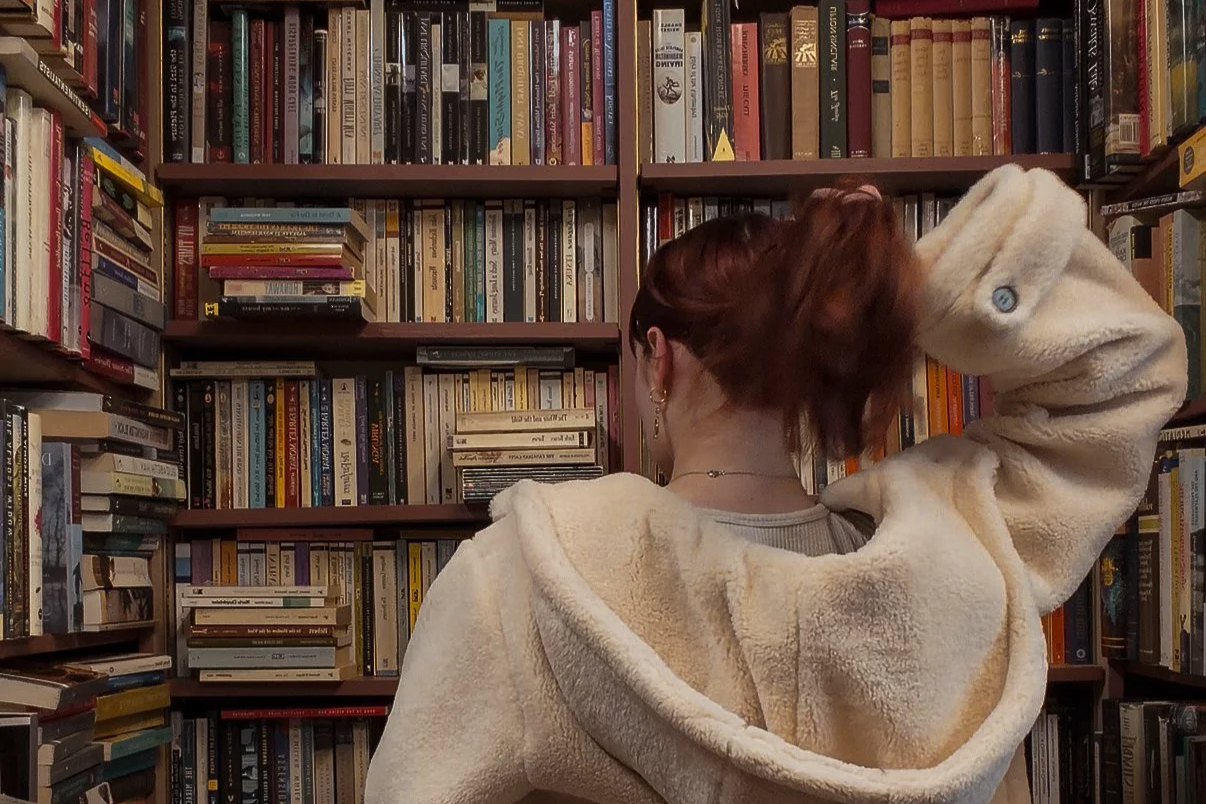 A person with red hair wearing a white fleece jacket standing in front of bookshelves filled with books, facing away from the camera.