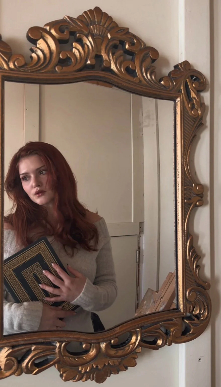 A woman with red hair taking a selfie in an ornate, carved gold mirror, holding a book with a decorative cover, standing in a room with a white door and cabinet behind her.