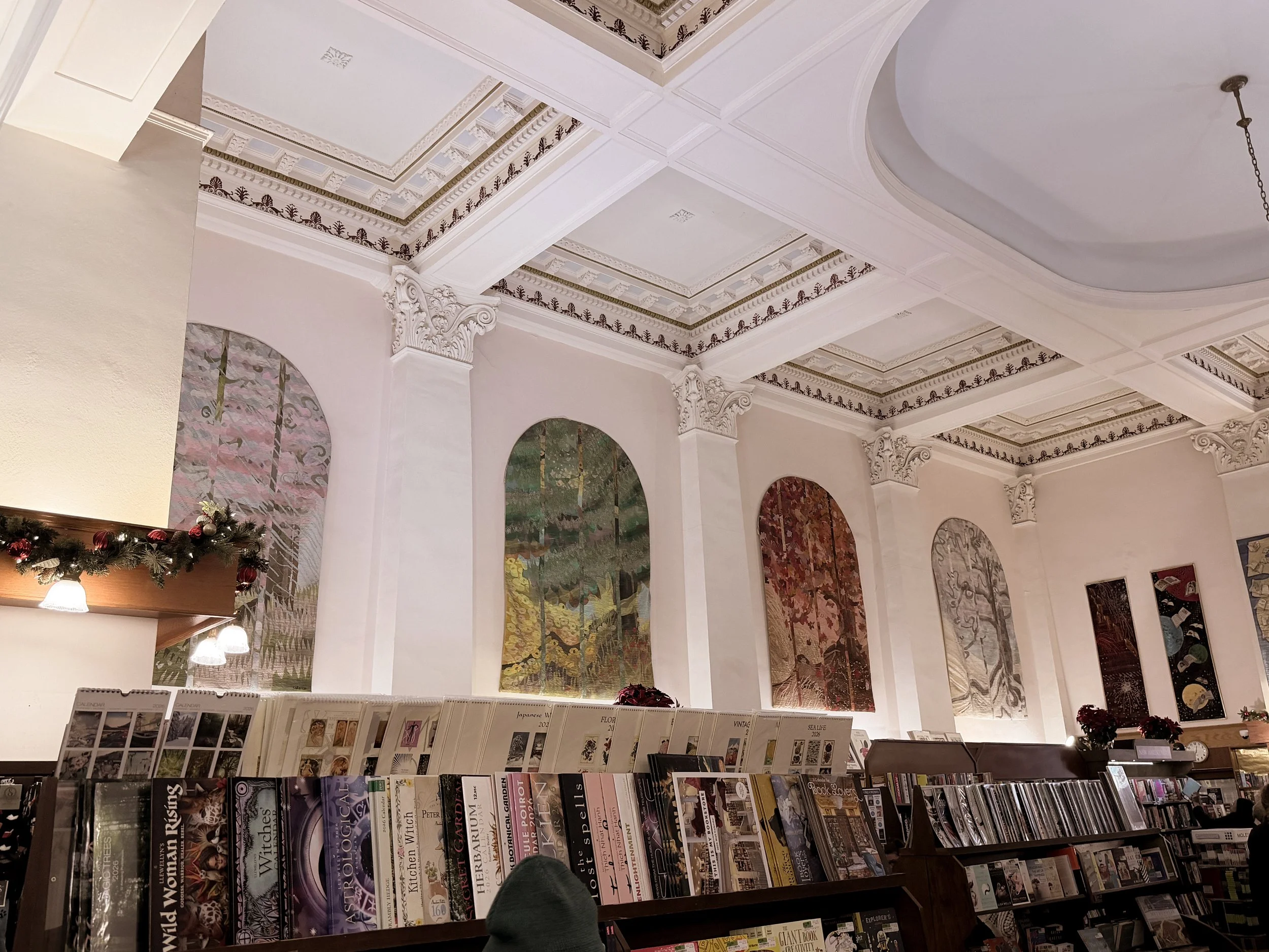 Inside a bookstore with high ceilings and large arched windows featuring artwork or posters, decorated with Christmas garland, and shelves of magazines and books.