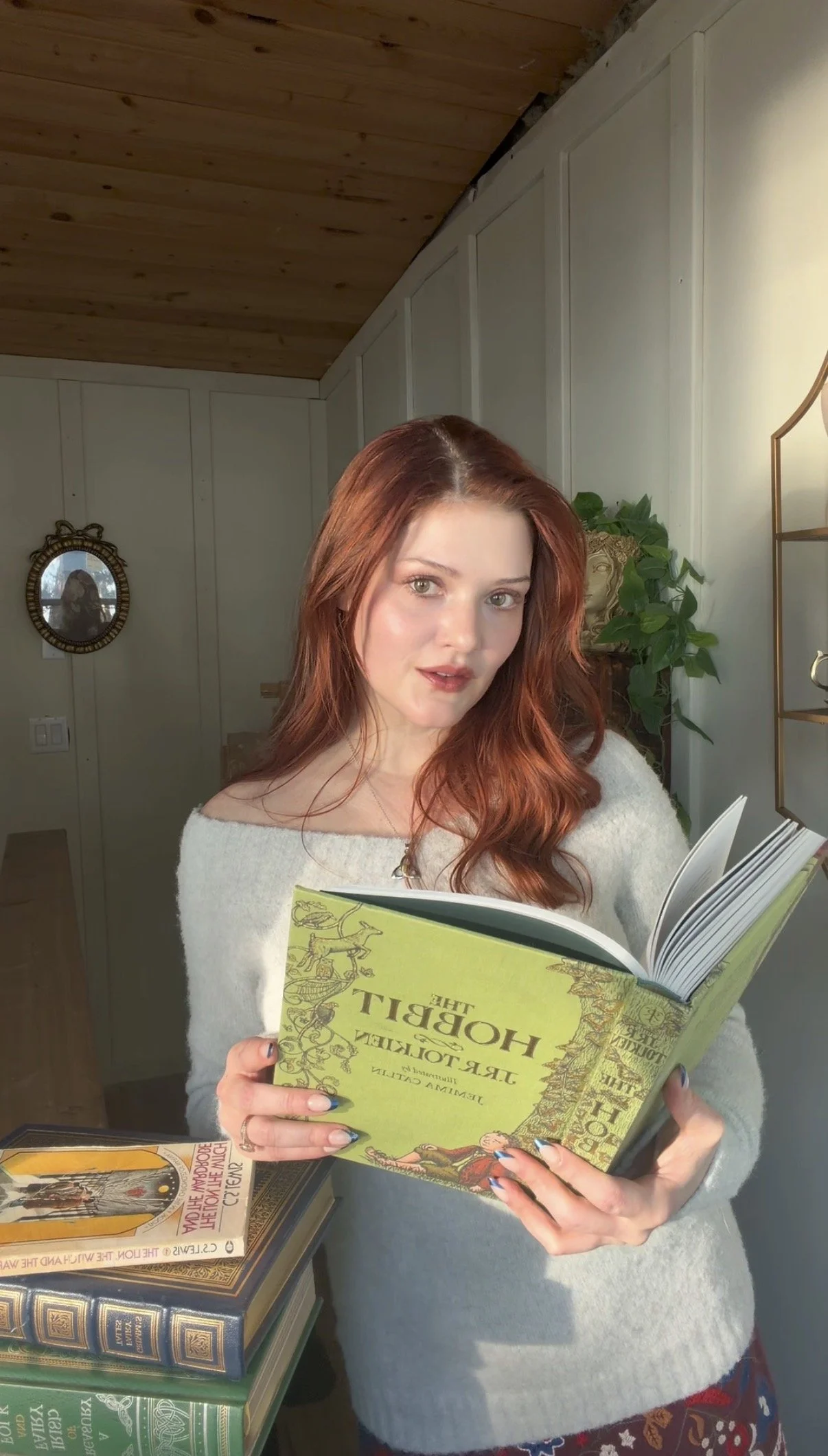 A woman with red hair is standing indoors, holding an open book titled "The Hobbit". She wears a light-colored sweater and features a gentle smile. There are several other books stacked on a table in front of her.