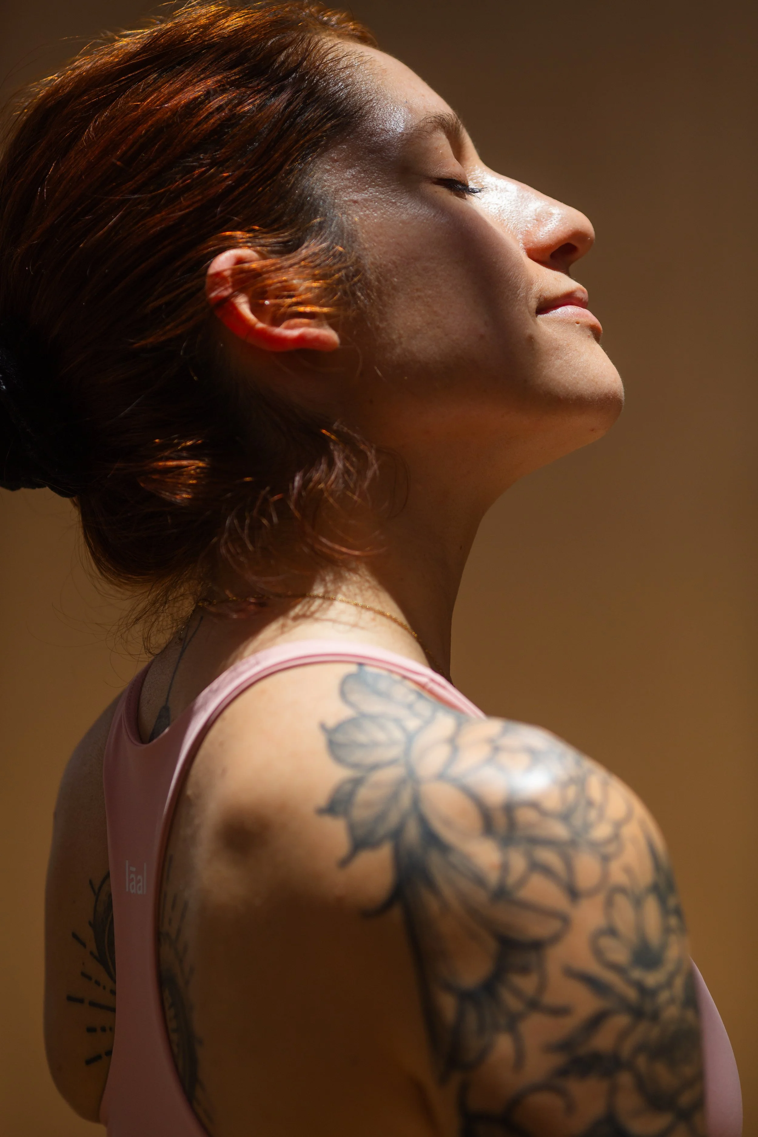 A woman with closed eyes, red hair, and tattoos on her shoulder, wearing a pink tank top.