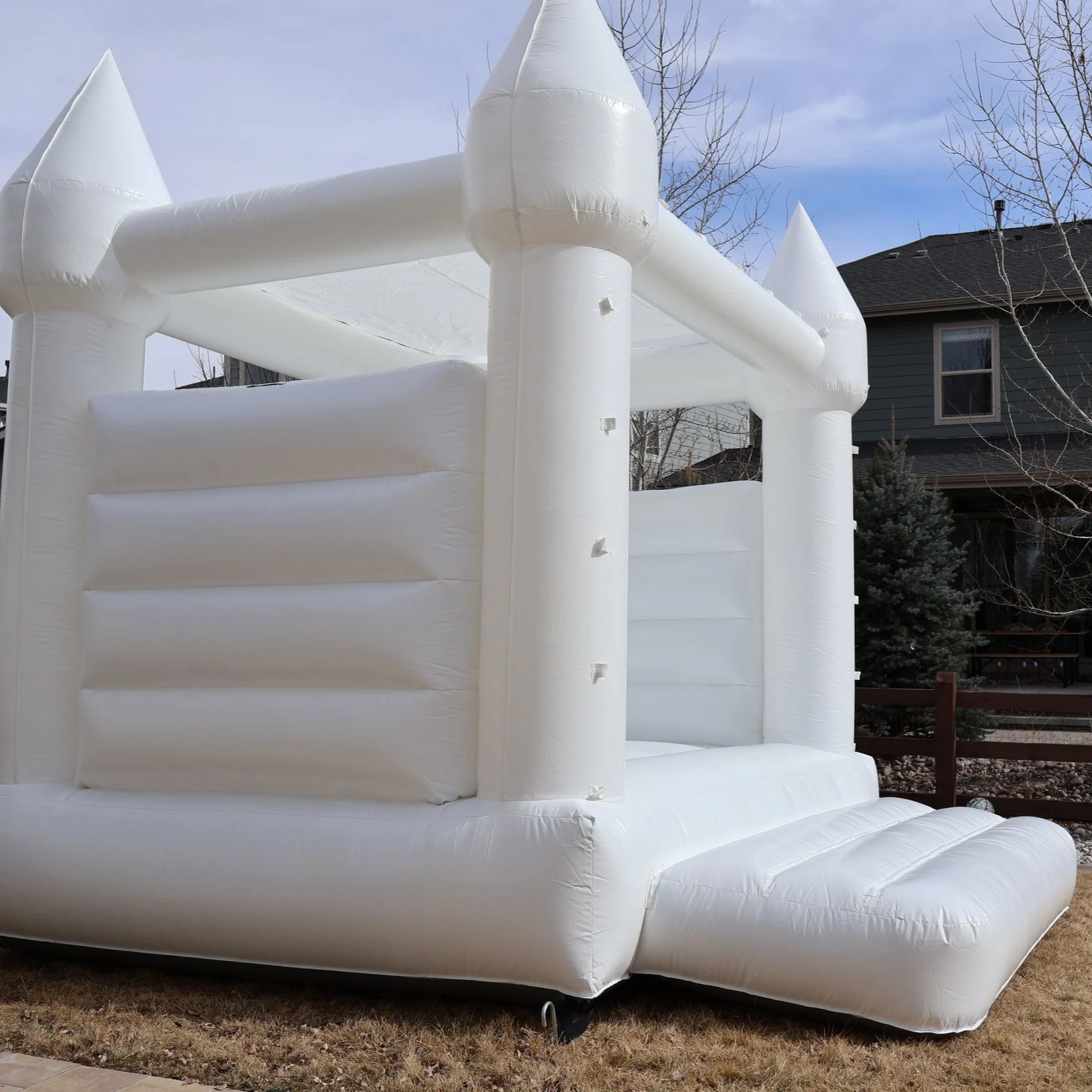 White inflatable castle for kids with towers, set up outdoors on grass in front of a house with a dark roof and trees.