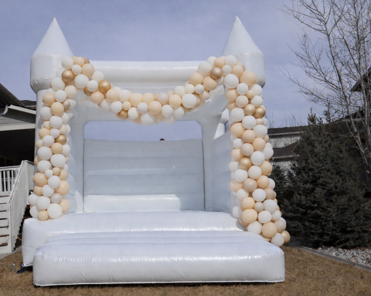 White inflatable castle decorated with beige, gold, and white balloons on its columns and around the top entrance, set outdoors with trees and a house in the background.