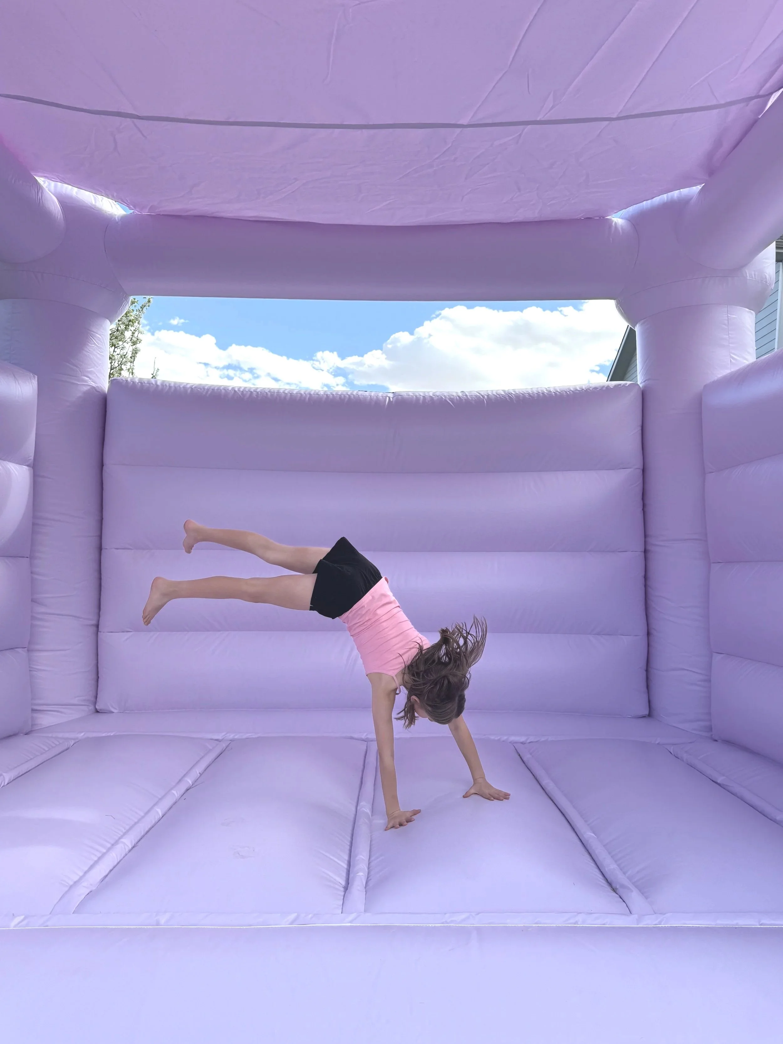 A young girl in a pink shirt and black shorts performing a handstand on an inflatable purple bounce house with a blue sky and white clouds in the background.