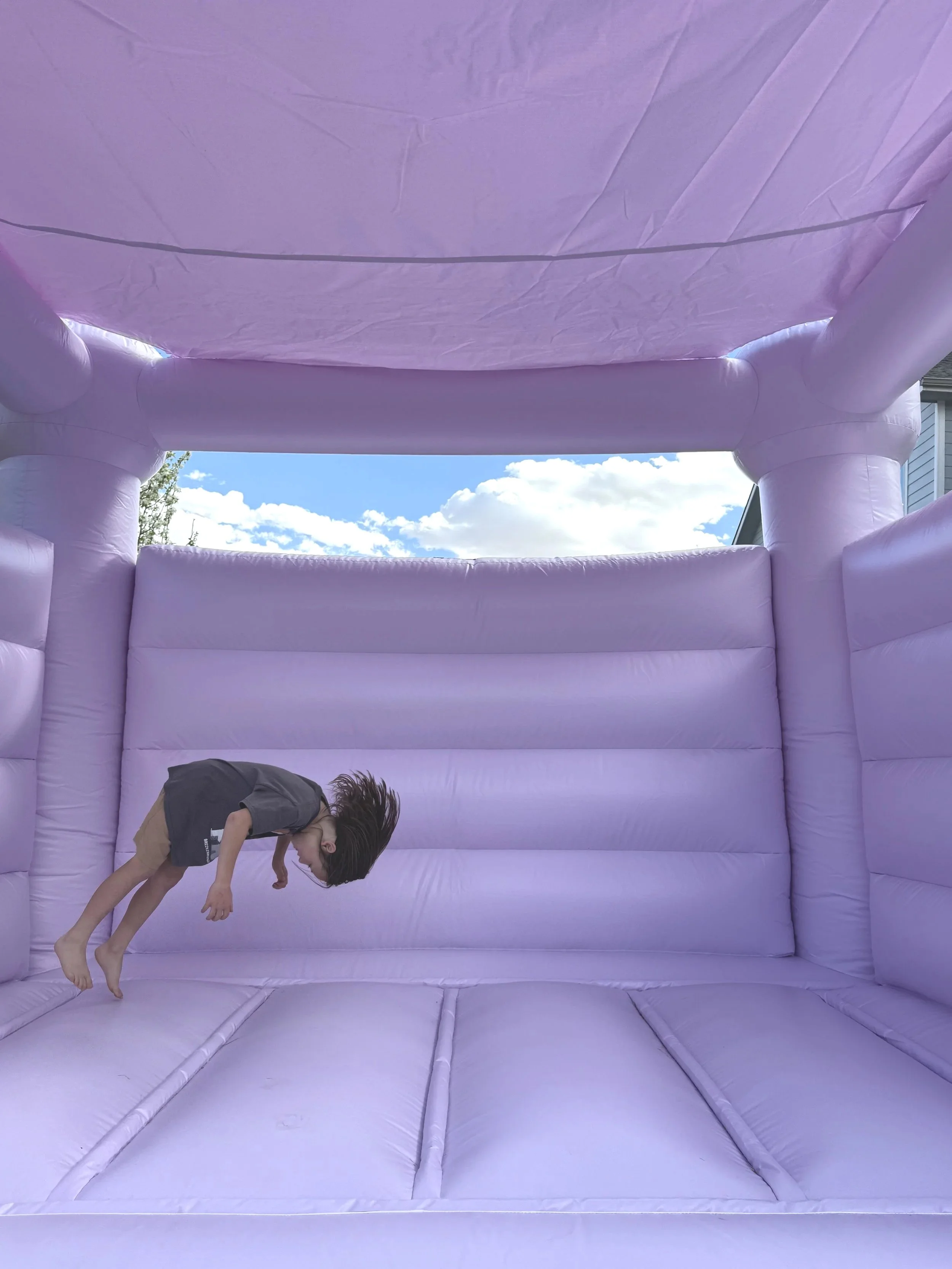 A child with long hair is bouncing on a purple inflatable bounce house outdoors, with blue sky and white clouds visible in the background.
