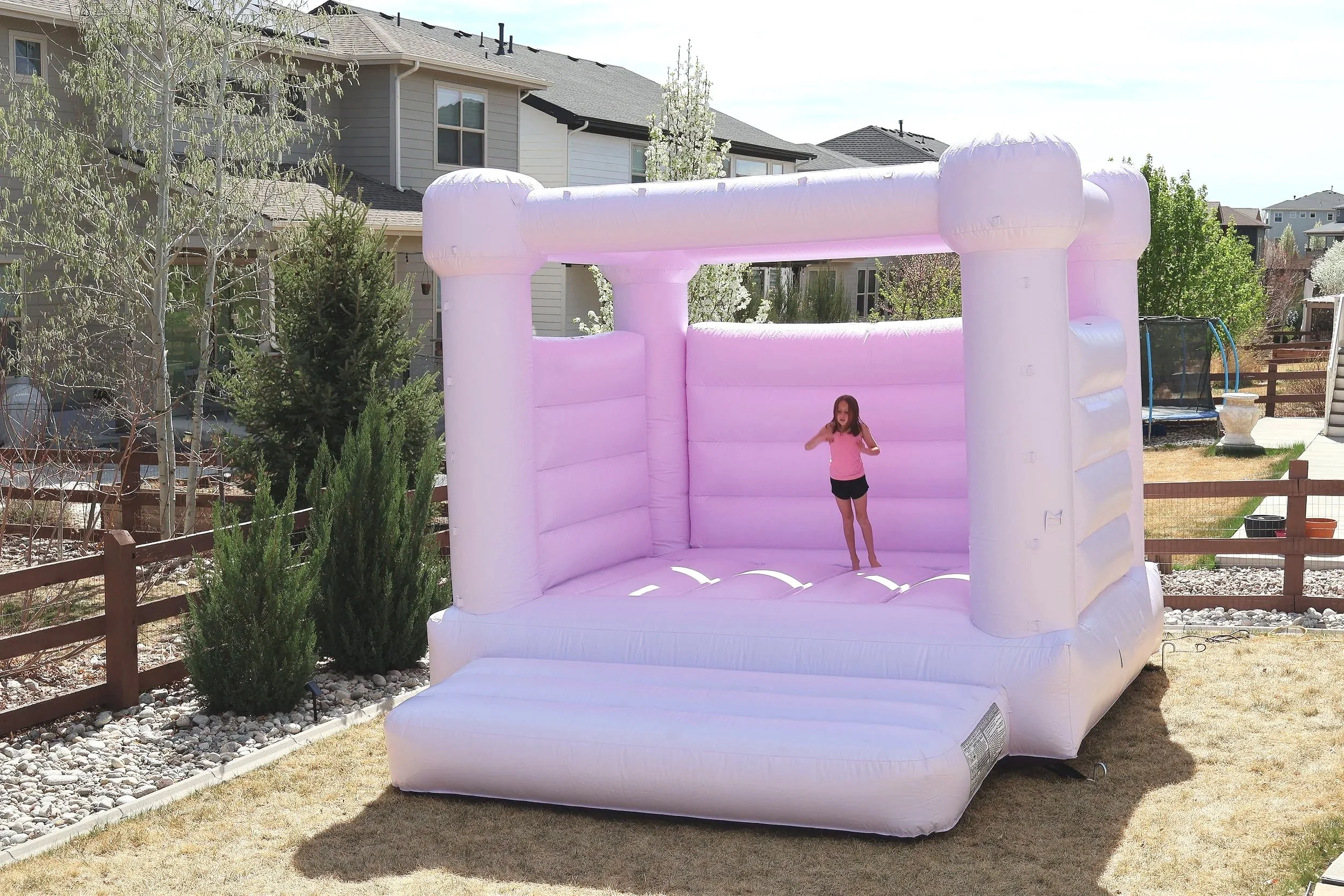 A young girl standing inside a pink and white inflatable bounce house in a suburban backyard, surrounded by trees, bushes, and houses.