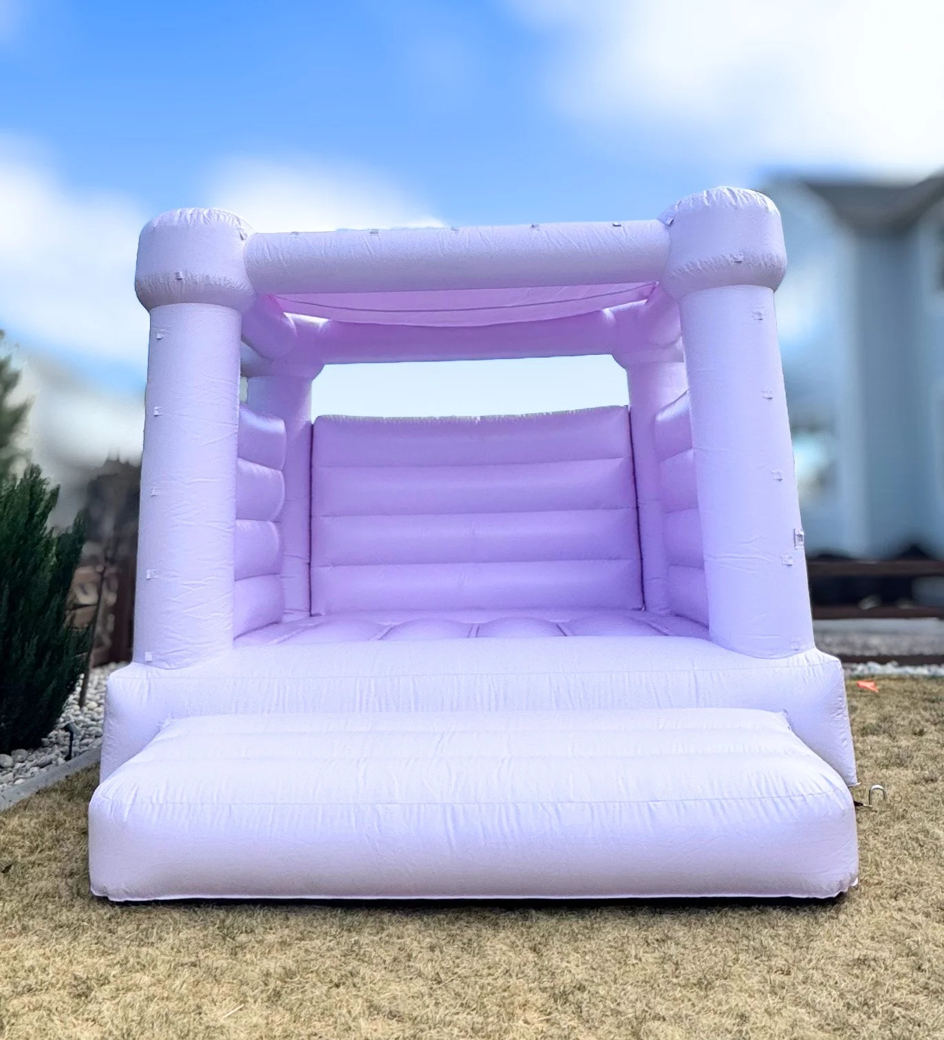 A white inflatable bounce house with a pinkish hue inside, set up outdoors on a grassy area, with a blue sky and some clouds in the background.