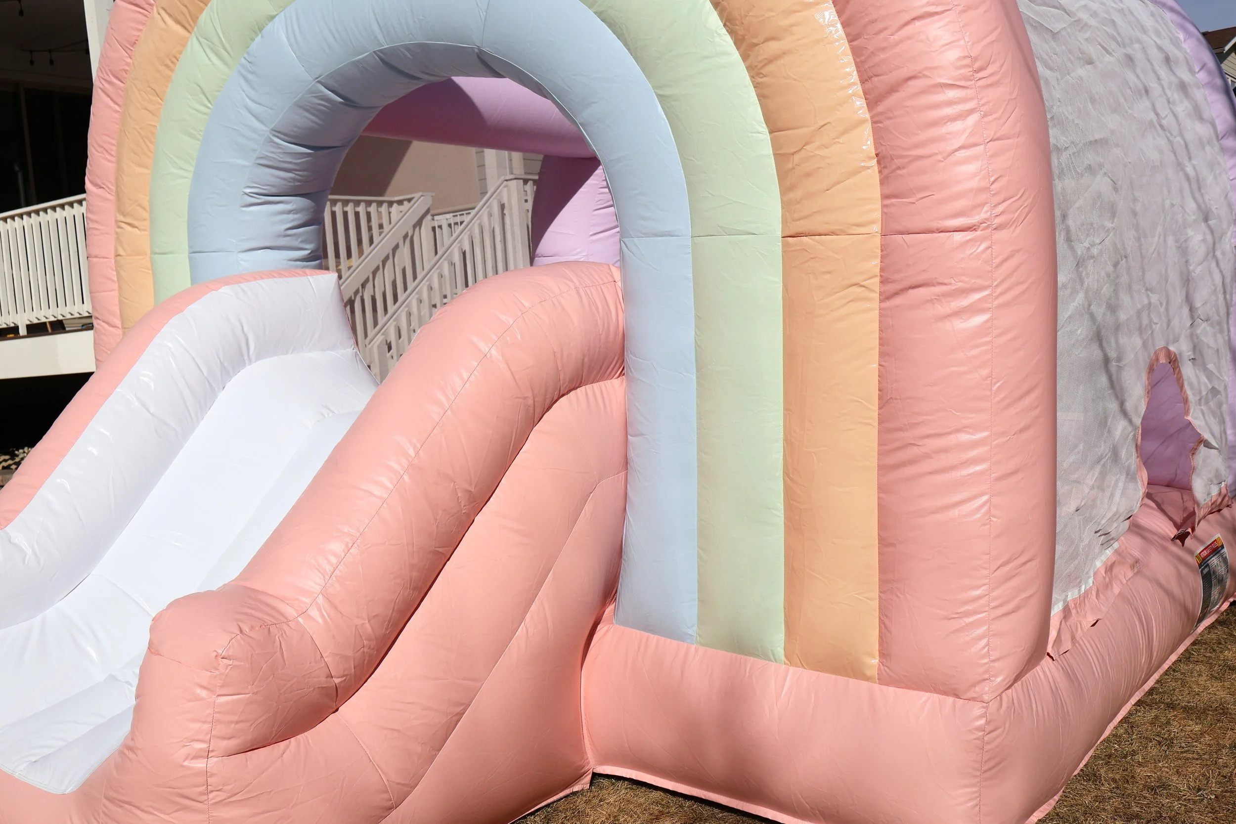 Colorful inflatable bounce house with a pink slide and rainbow arch entrance, set up outdoors on grass.