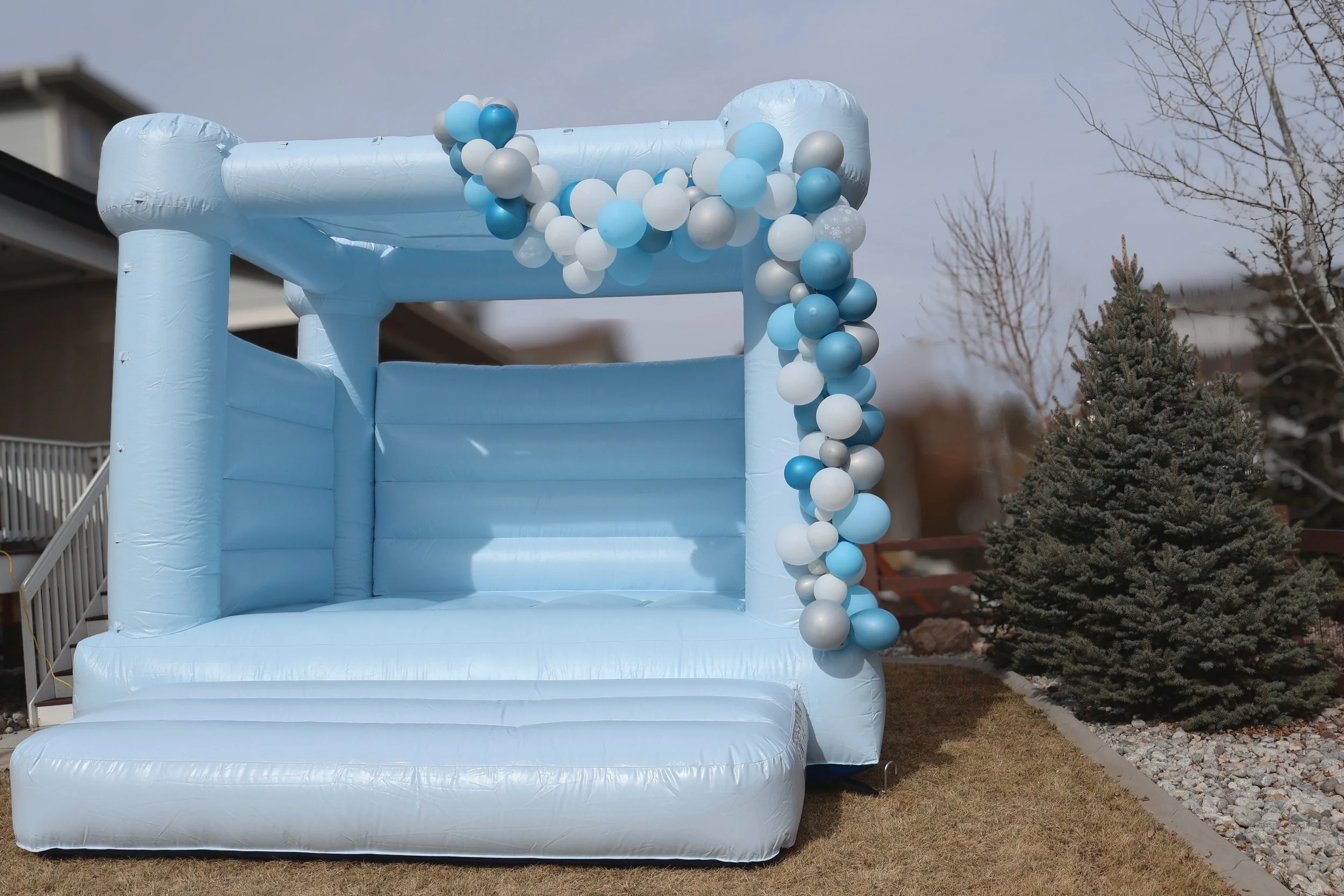 Blue inflatable bounce house with white, blue, and gray balloons attached, set up outdoors on a grassy yard near a pine tree and residential house.