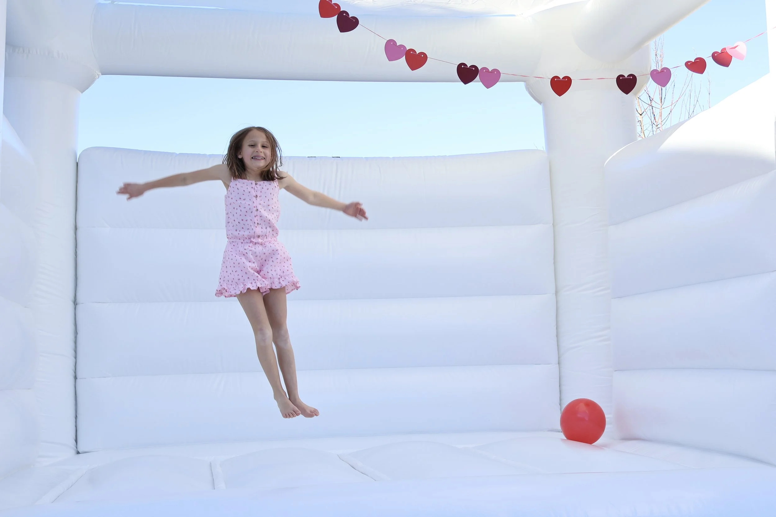 Young girl in pink pajamas jumping on a white inflatable bounce house with a red ball inside, decorated with heart-shaped garland against a blue sky.