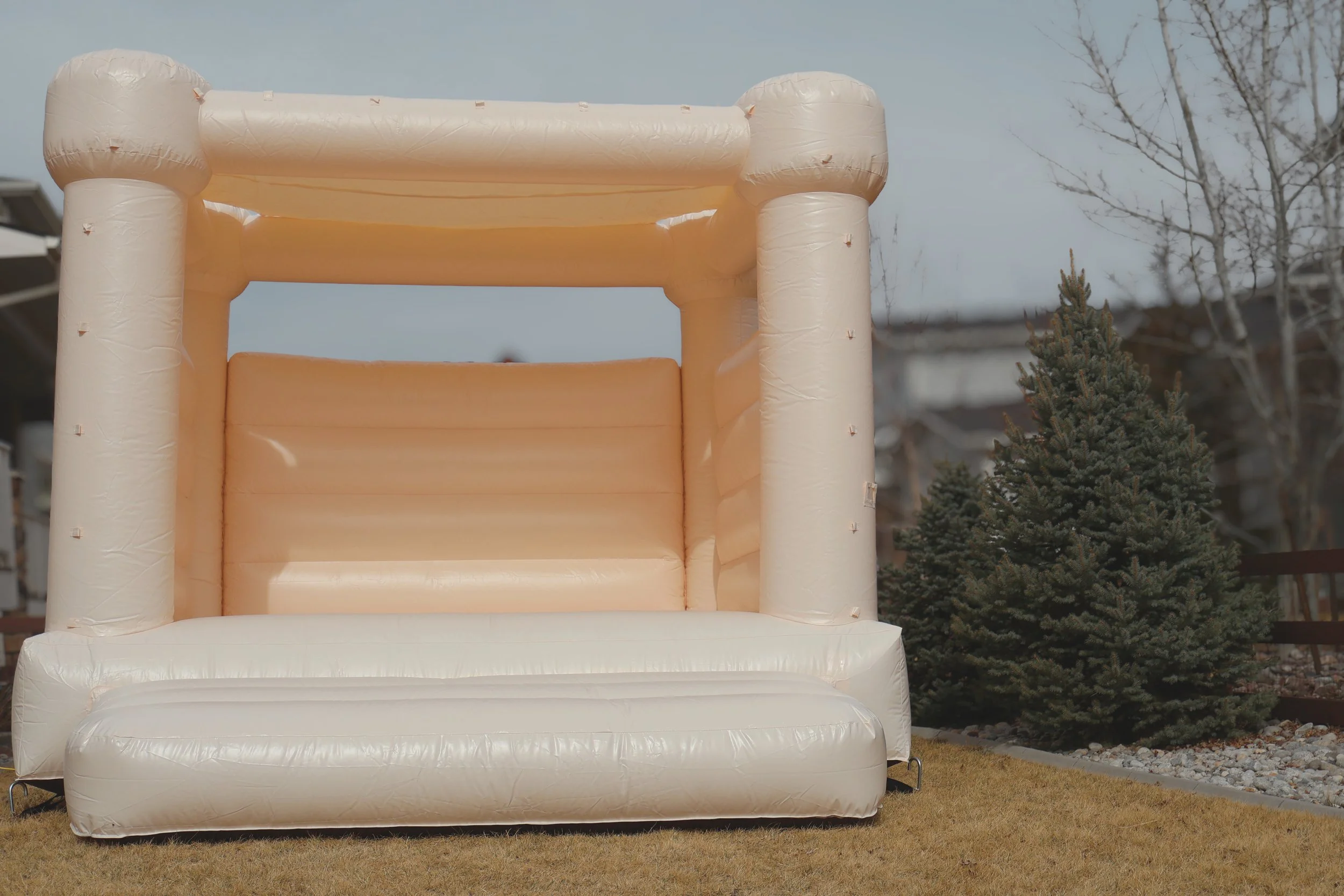 Inflatable outdoor bounce house with beige and peach colors on a grassy yard, near a small evergreen tree and some leafless trees, under a cloudy sky.