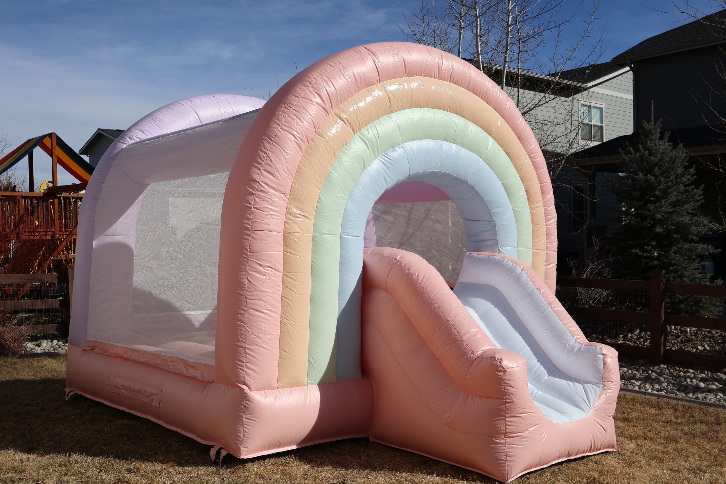 Inflatable bounce house with rainbow design, slide, pink, yellow, green, blue, purple, set up on a lawn in a residential backyard.