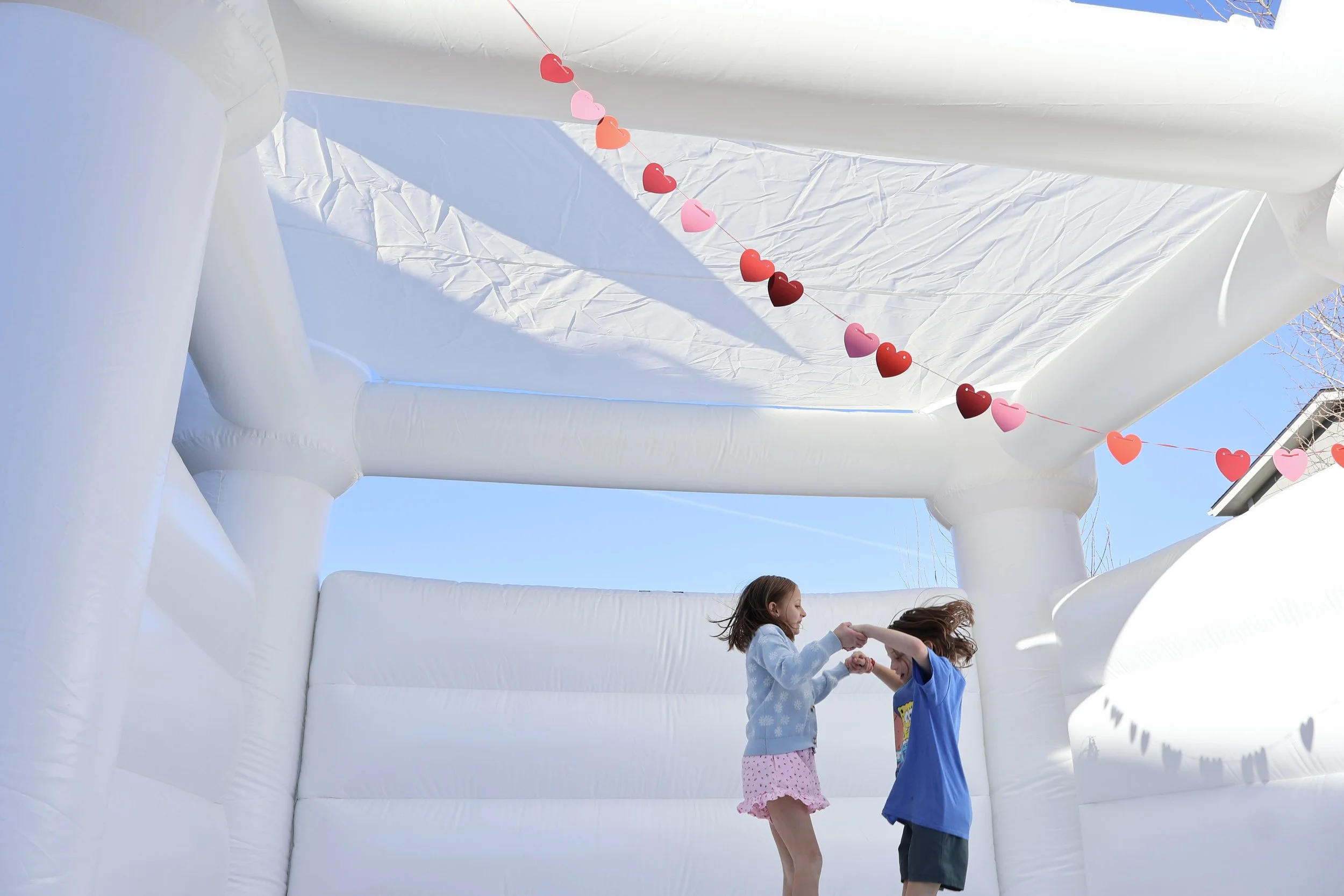 Two children, a girl and a boy, playing and holding hands inside a white bounce house with a blue sky overhead. Decorative heart-shaped garland is hanging inside the bounce house.