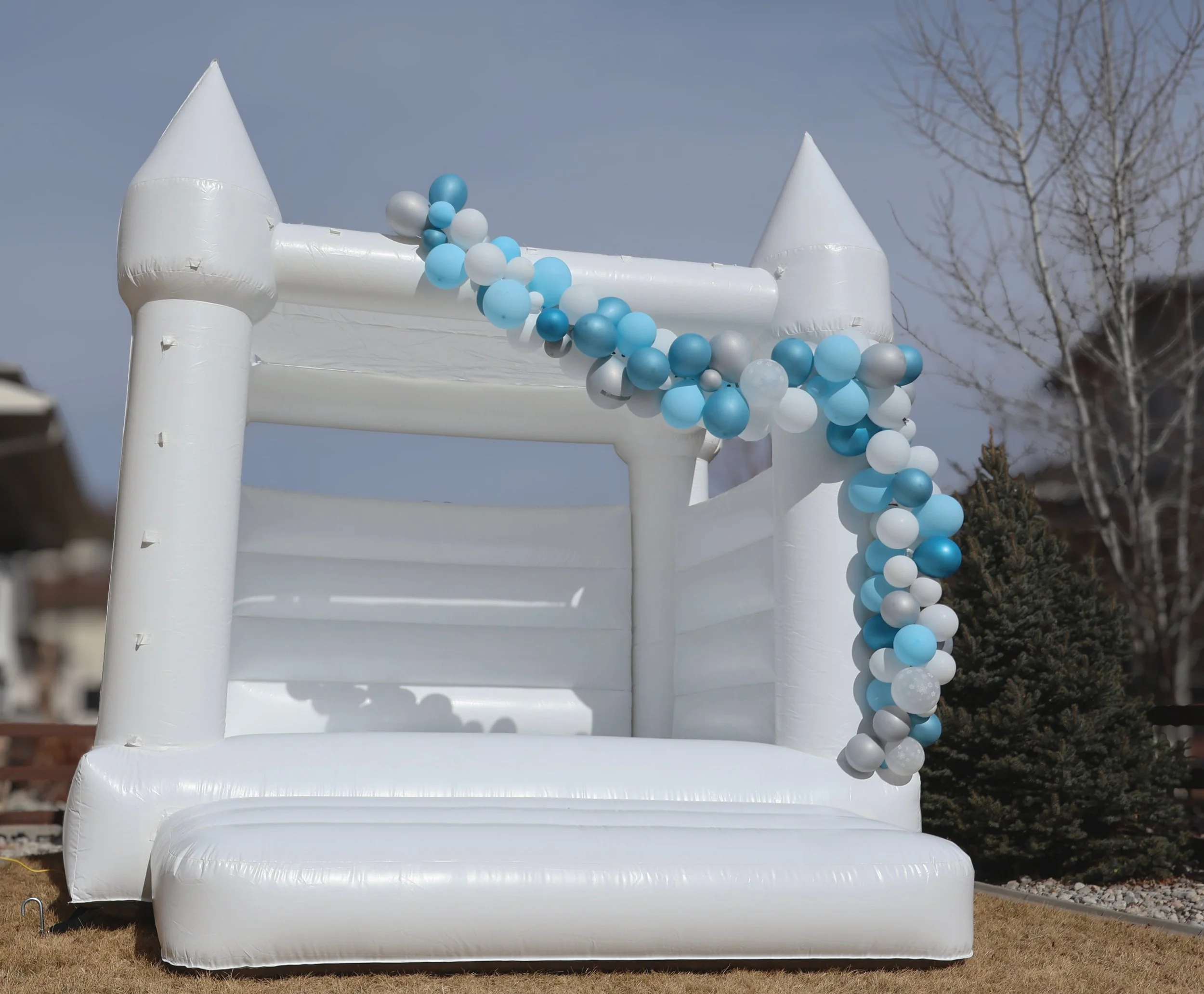 White inflatable castle decorated with blue, white, and silver balloons in an outdoor yard with trees and houses in the background.