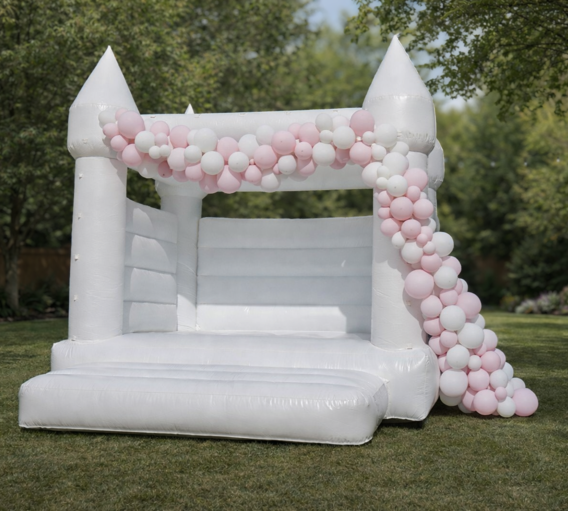 White inflatable castle with pink and white balloons decorating the archway, set up outdoors on grass grass with trees in the background.
