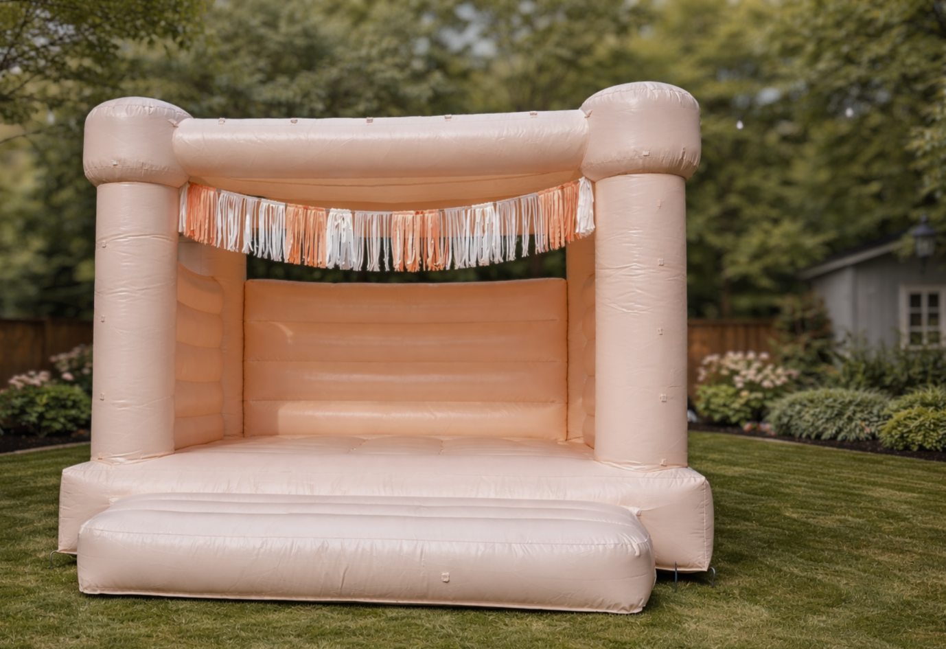 Pink inflatable bounce house with a decorative banner, set up in a backyard with green grass, trees, and a small shed in the background.
