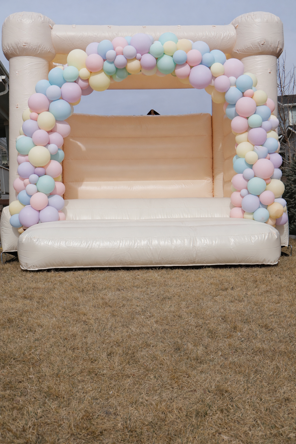 Inflatable beige bounce house with pastel-colored balloon decorations in pink, blue, yellow, purple, and green around the entrance, set outdoors on grass.