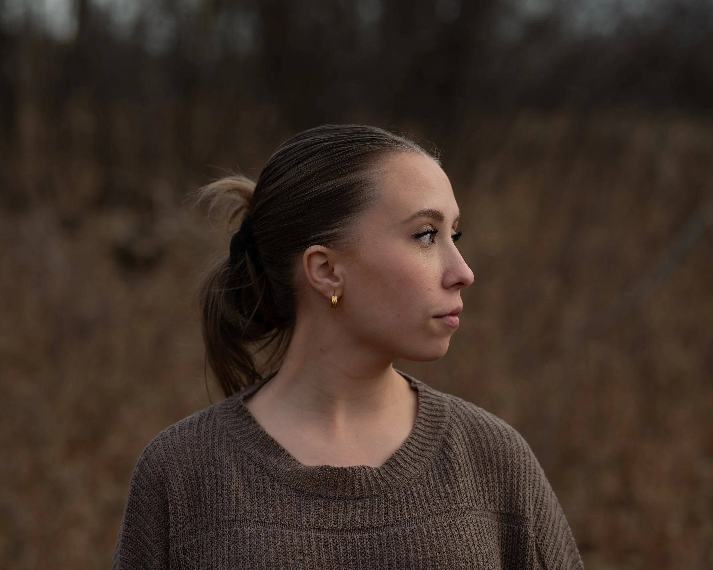 Profile of a young woman with brown hair tied back, wearing a dark brown sweater and small gold hoop earrings, outdoors with a blurred background of trees and grass.