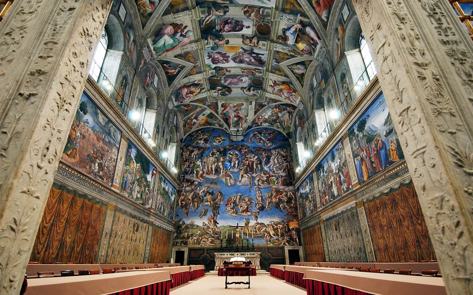 A large, ornate church or chapel interior with high vaulted ceilings covered in religious paintings, including scenes with numerous figures, angels, and biblical themes. There are long tables with red tablecloths on either side of the central aisle, and a small altar at the front with candles and a cross.