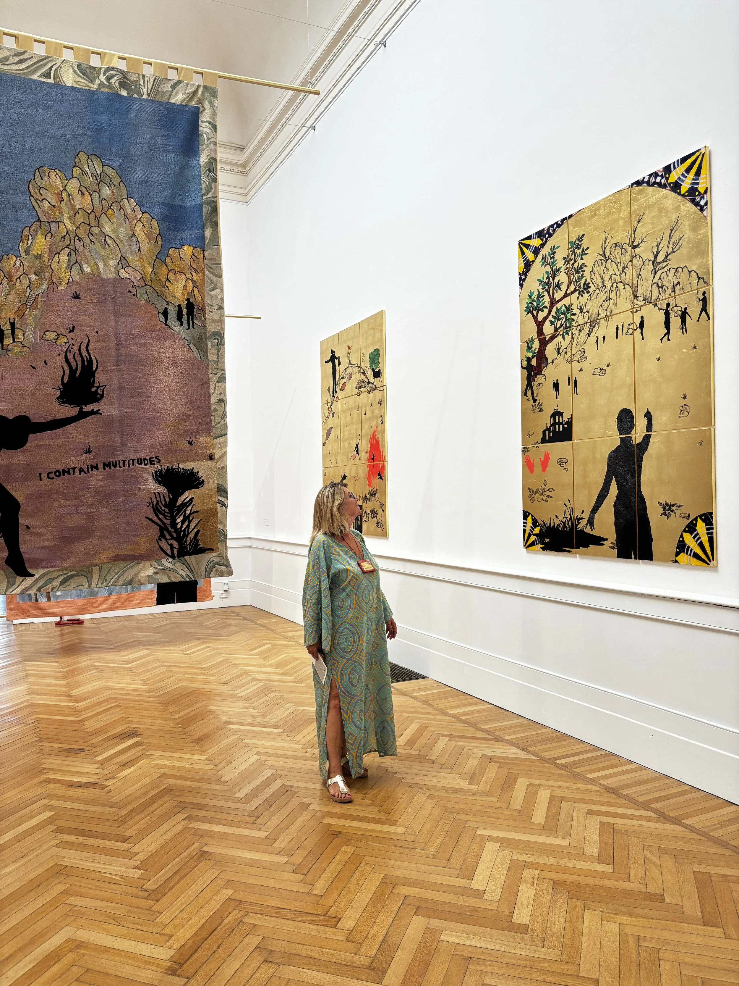 A woman in a long, colorful dress with a slit, white sandals, and blonde hair viewing art in a gallery with wooden herringbone parquet floor, white walls, and hanging artworks.