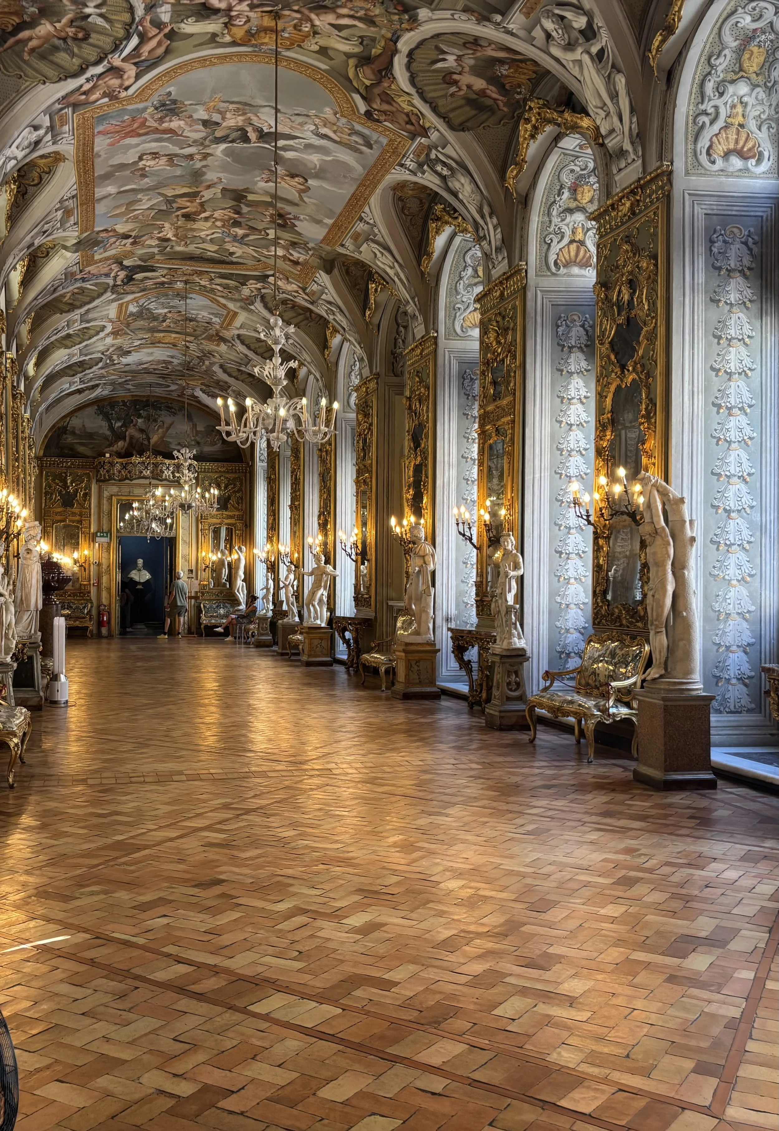 The image shows an ornate historic gallery with high ceiling, decorated with detailed frescoes and gilded moldings. The room has gold-framed mirrors, chandeliers hanging from the ceiling, and rows of classical sculptures along the walls. The floor is made of arranged bricks, and a few visitors are seen exploring the space.