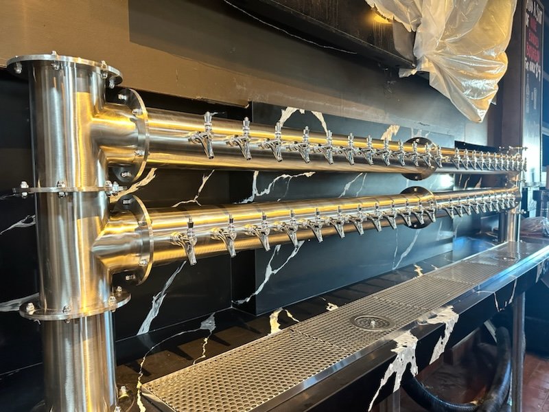 Two stainless steel beer taps mounted on a black marble wall with white veins, set above a metal drip tray.