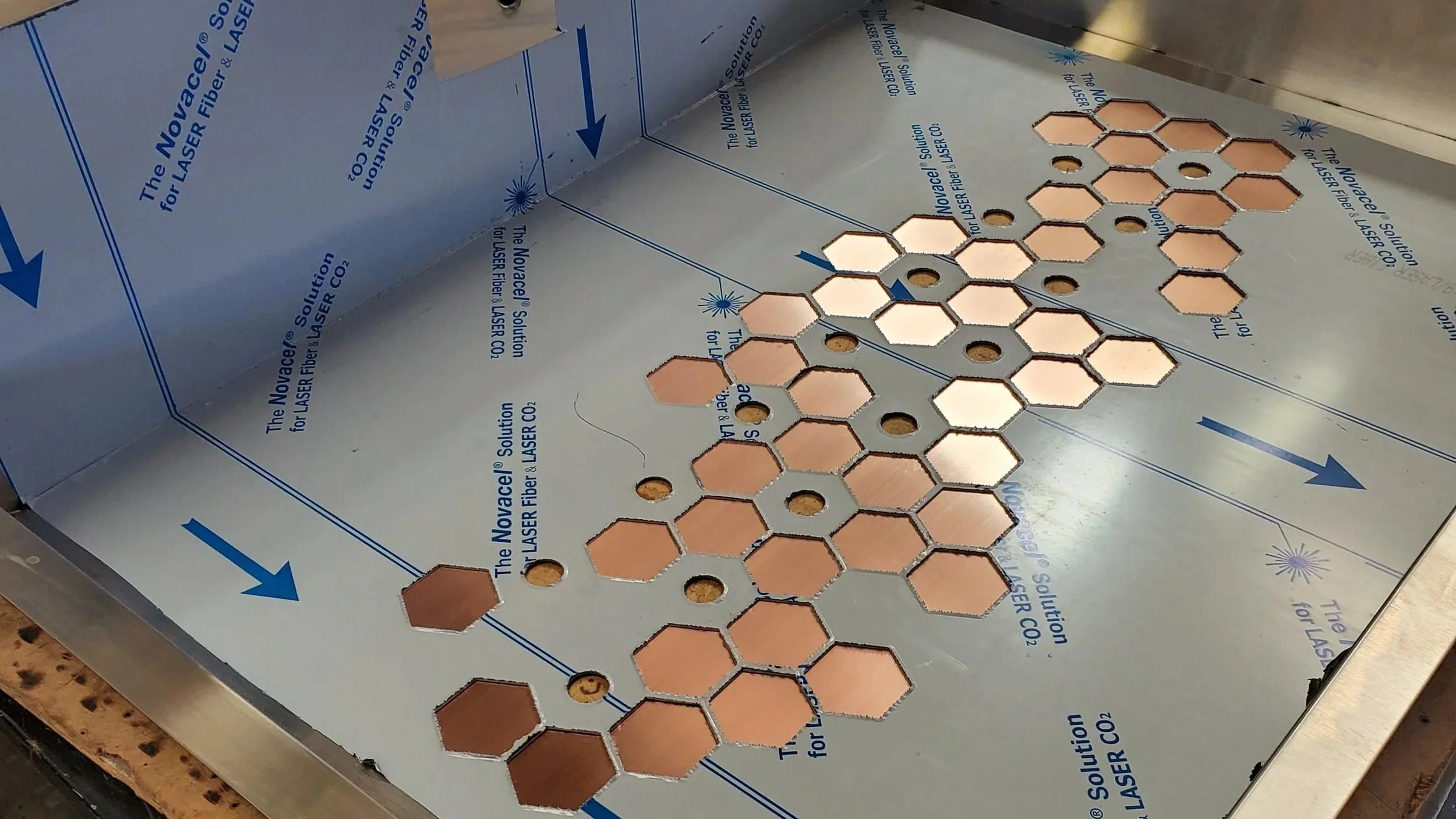 A metal surface with hexagonal copper pieces and holes, covered with protective blue tape marked 'The Novacel Solution for LASER Fiber LASER CO2' along with arrows and lines for alignment.