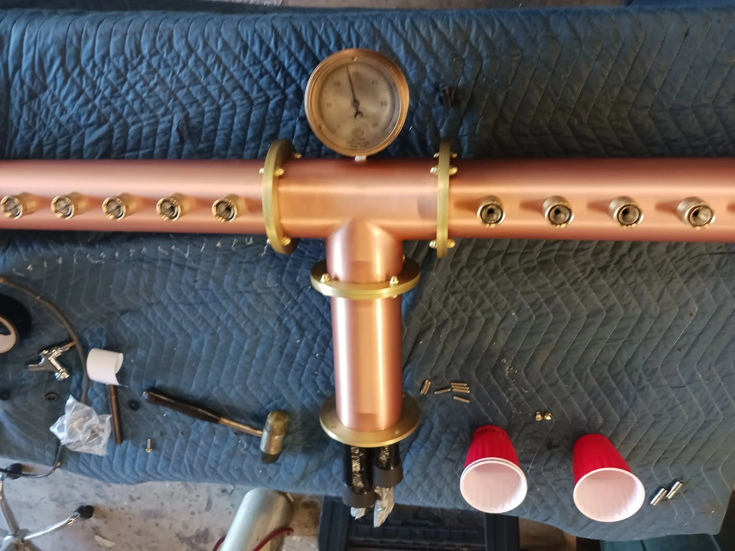 Copper and brass structure of a distillation apparatus with gauges, tools, and plastic cups on a quilted blue surface.