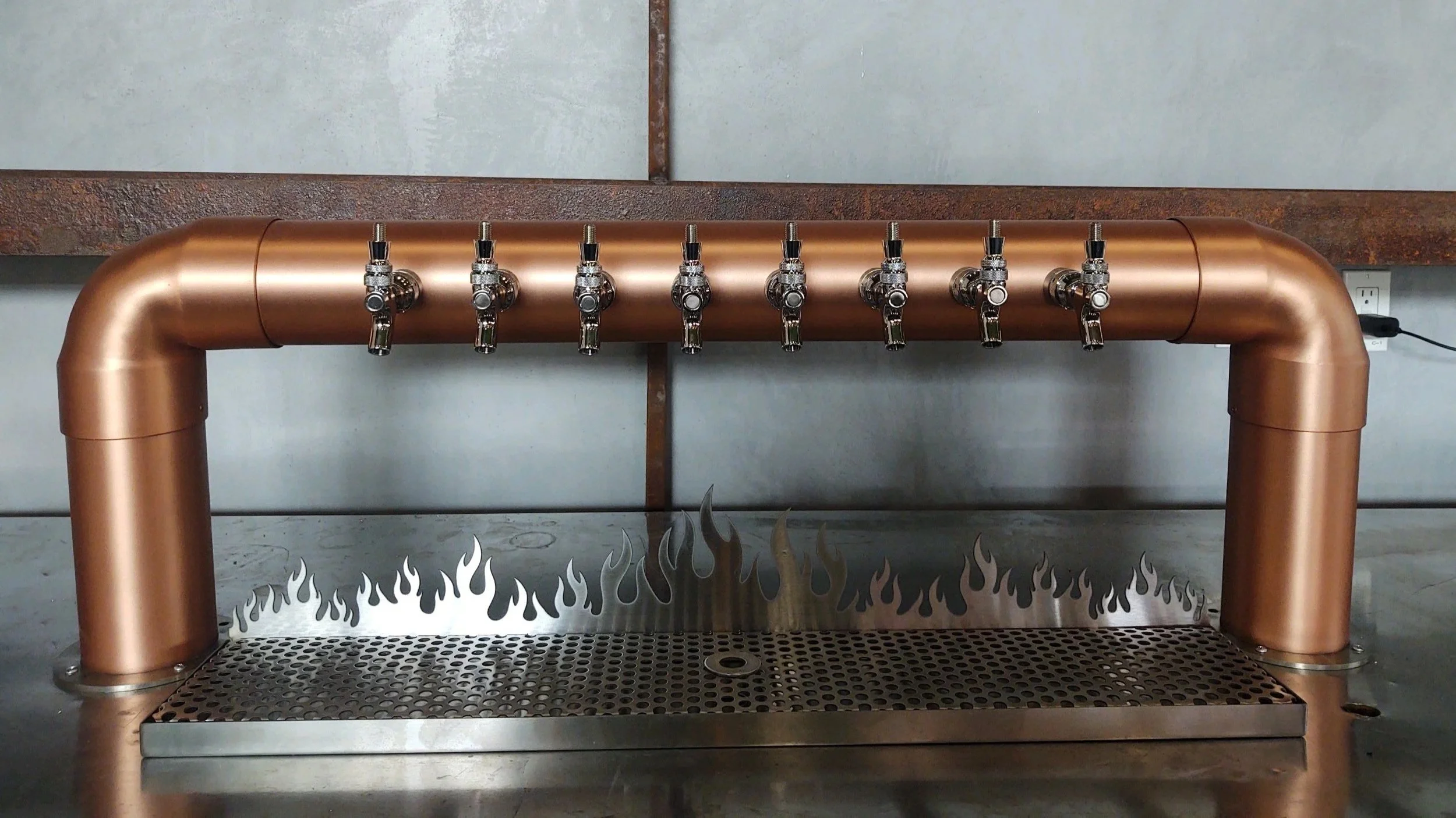 Copper beer tap system with seven faucets, decorative flame design on the backsplash, and a metal drip tray.