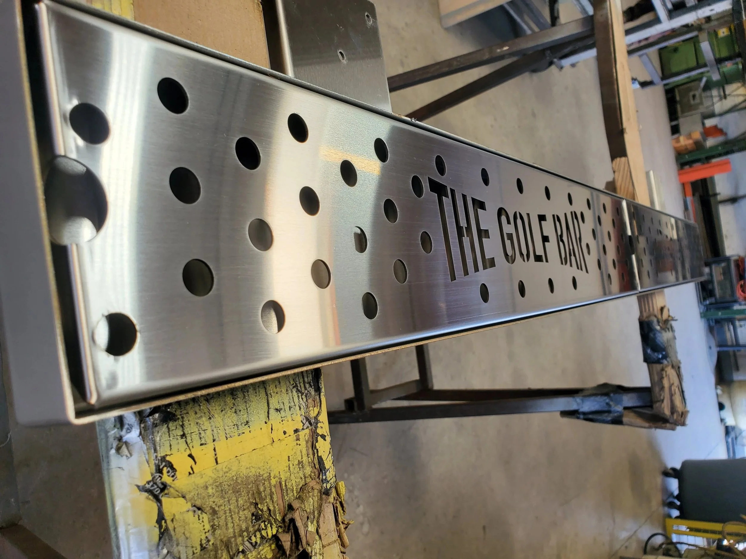A metallic sign with holes and the engraved text "THE GOLF BANK" in a workshop setting.